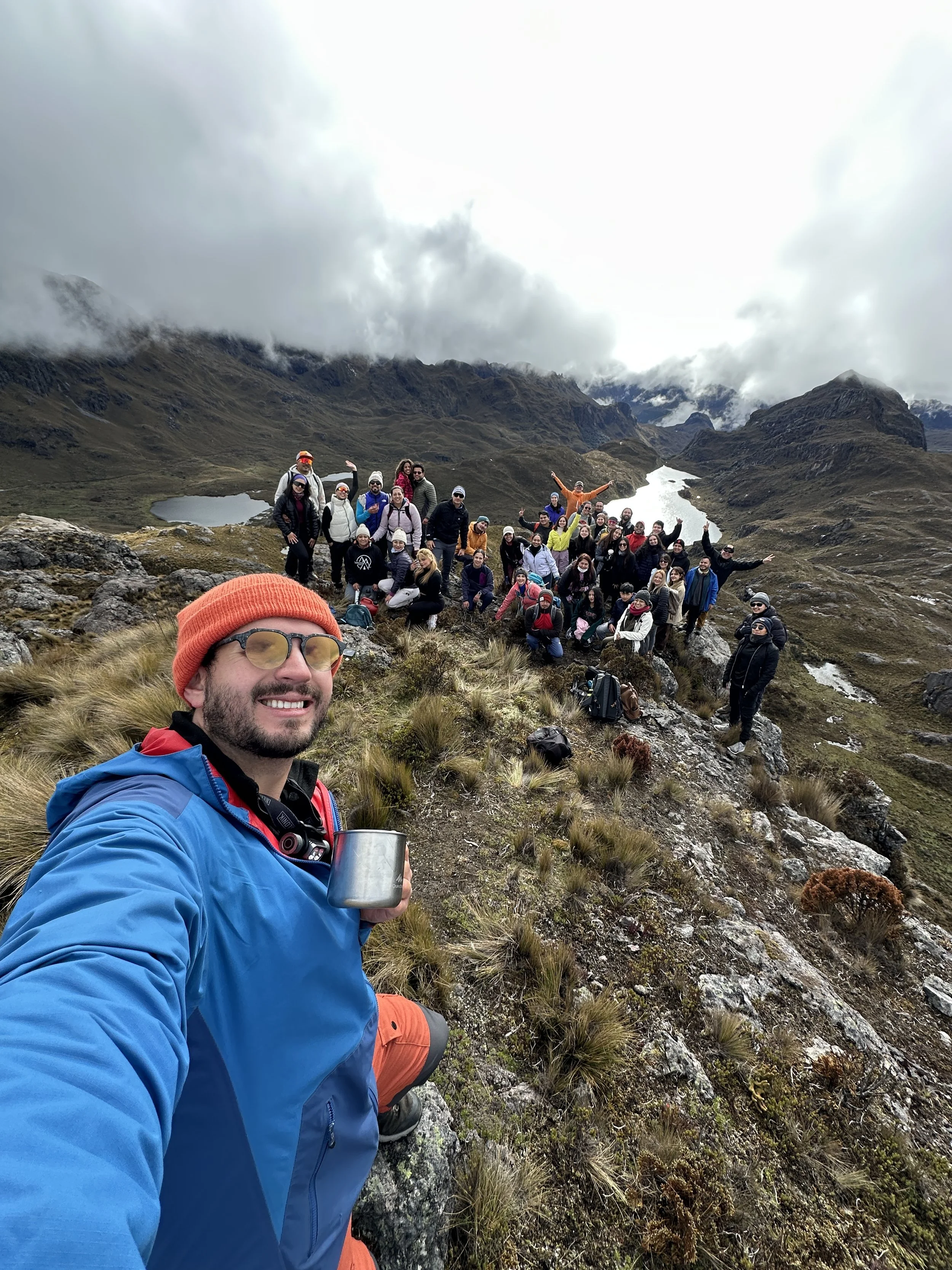 Grupo de personas haciendo senderismo en una región montañosa con lagunas y nubes en el cielo, una persona en primer plano con ropa azul y gorro anaranjado toma una selfie.
