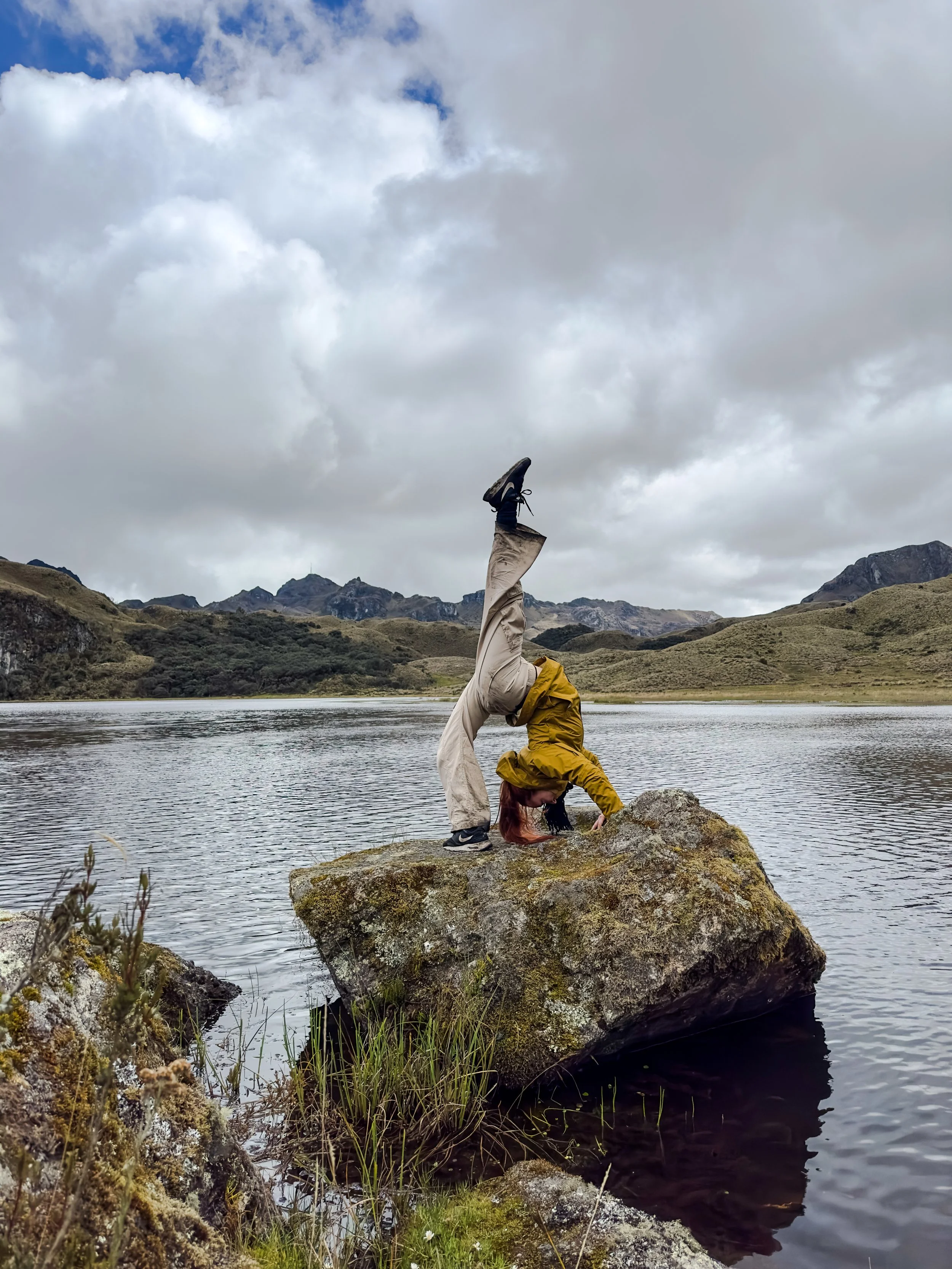 Persona haciendo una pose de yoga en una roca junto a un lago, con montañas y cielo nublado en el fondo.