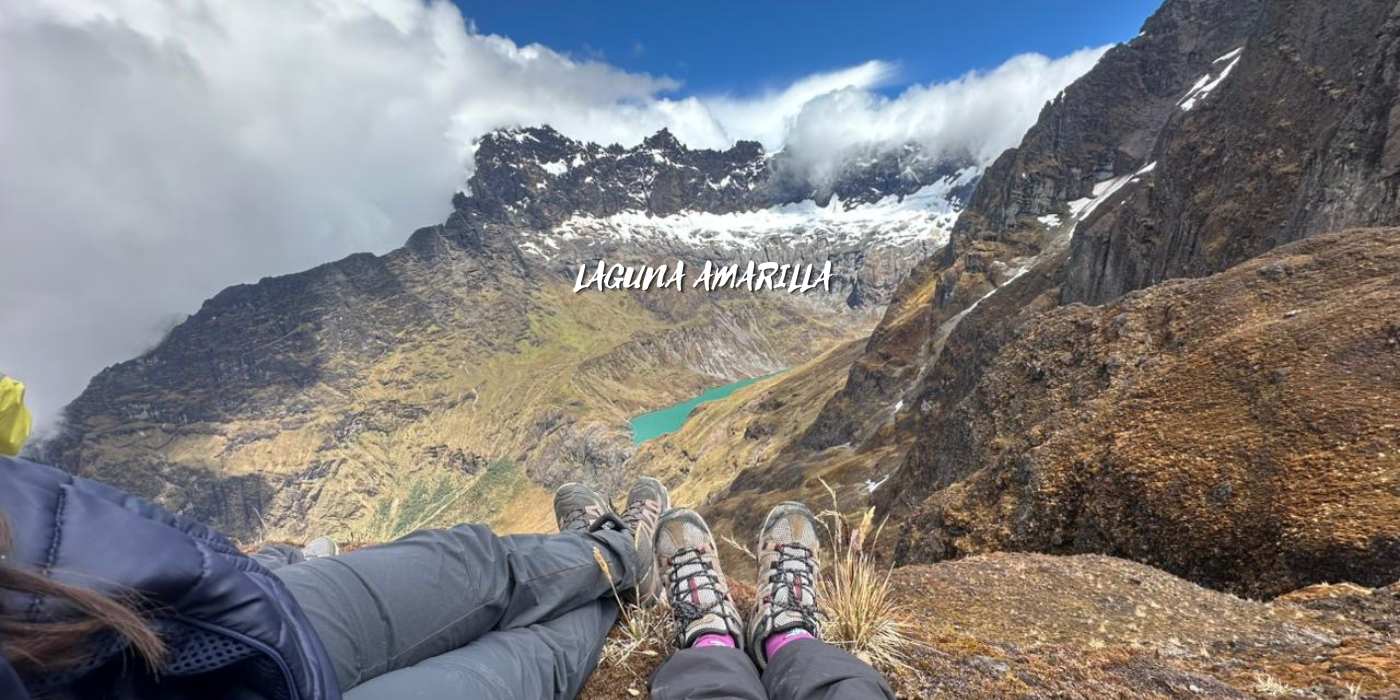 Personas descansando en la cima de una montaña con vista a una laguna amarilla en un paisaje montañoso con picos y nubes.