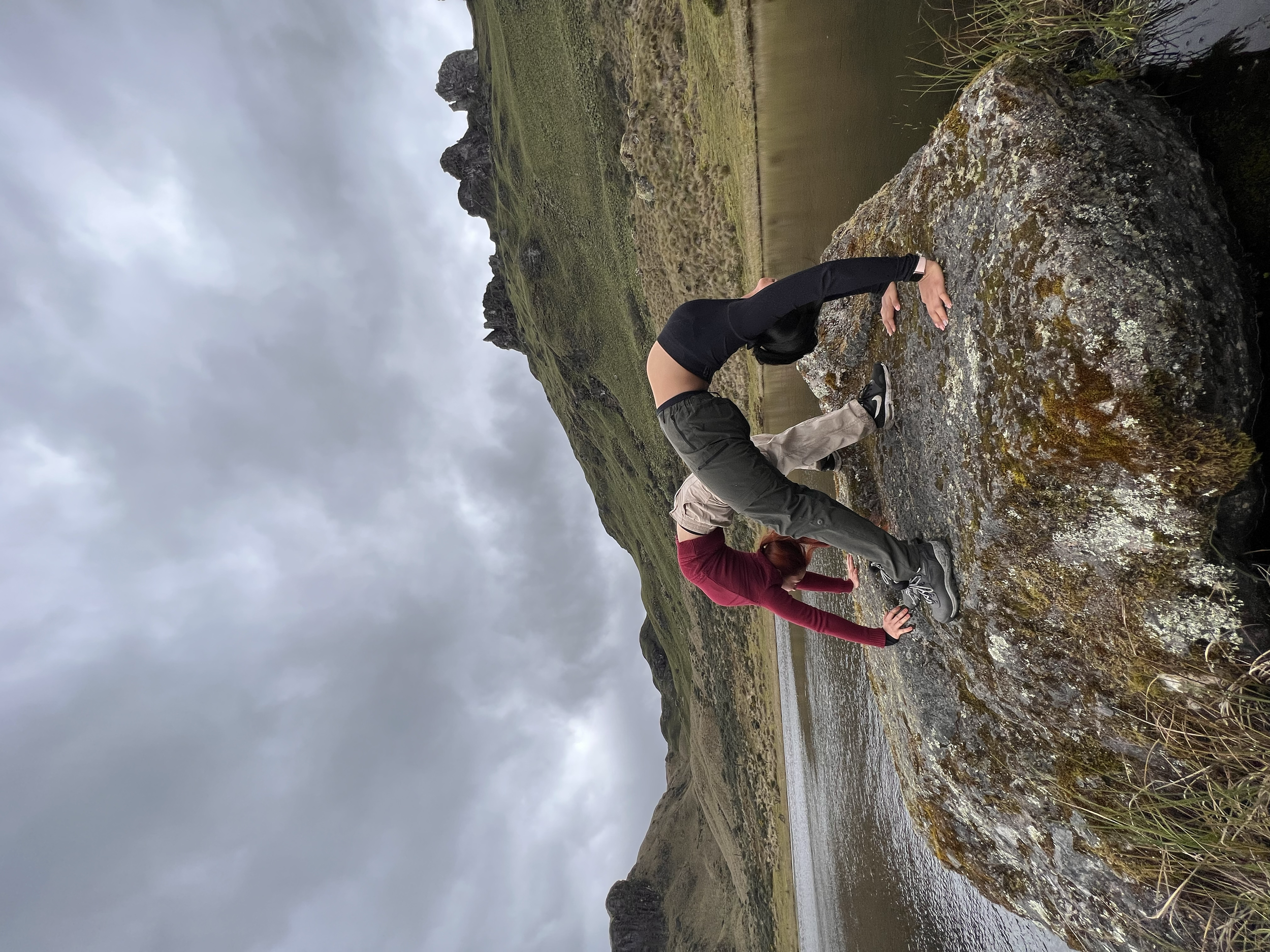 Dos personas en ropa de senderismo explorando una roca cerca de un río bajo un cielo nublado en un paisaje natural montañoso.