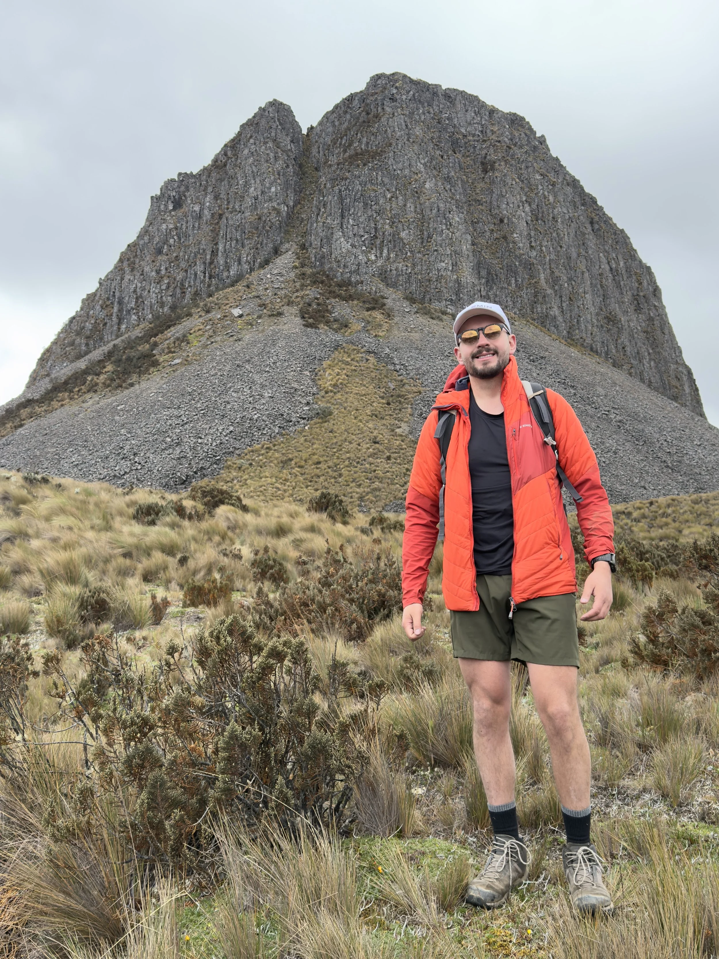 Persona con ropa de senderismo en un terreno de hierba y arbustos, con una montaña o roca grande al fondo bajo un cielo nublado.