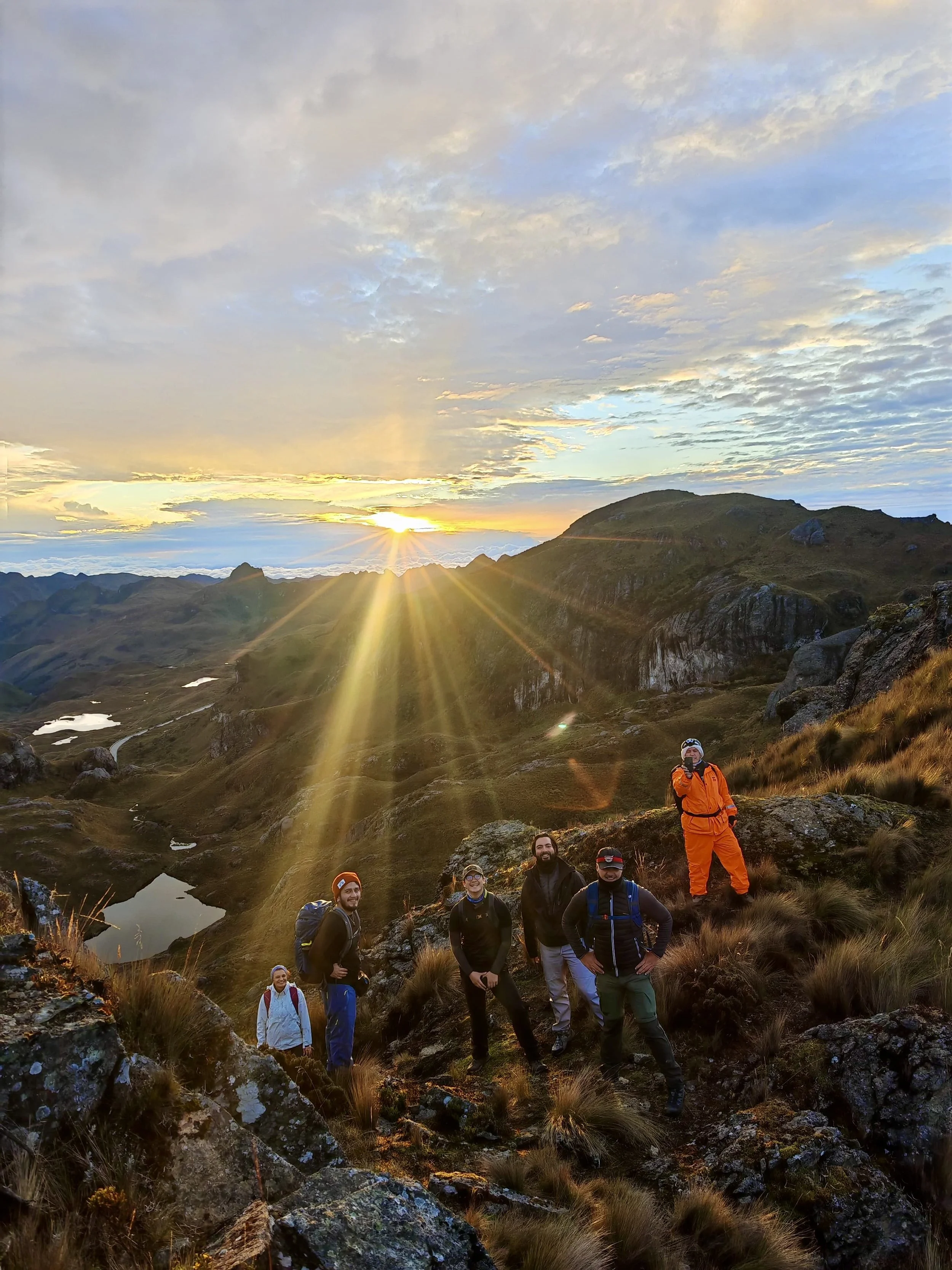 Un grupo de cinco excursionistas en una montaña al atardecer, viendo el sol ponerse en el horizonte con nubes y un paisaje montañoso
