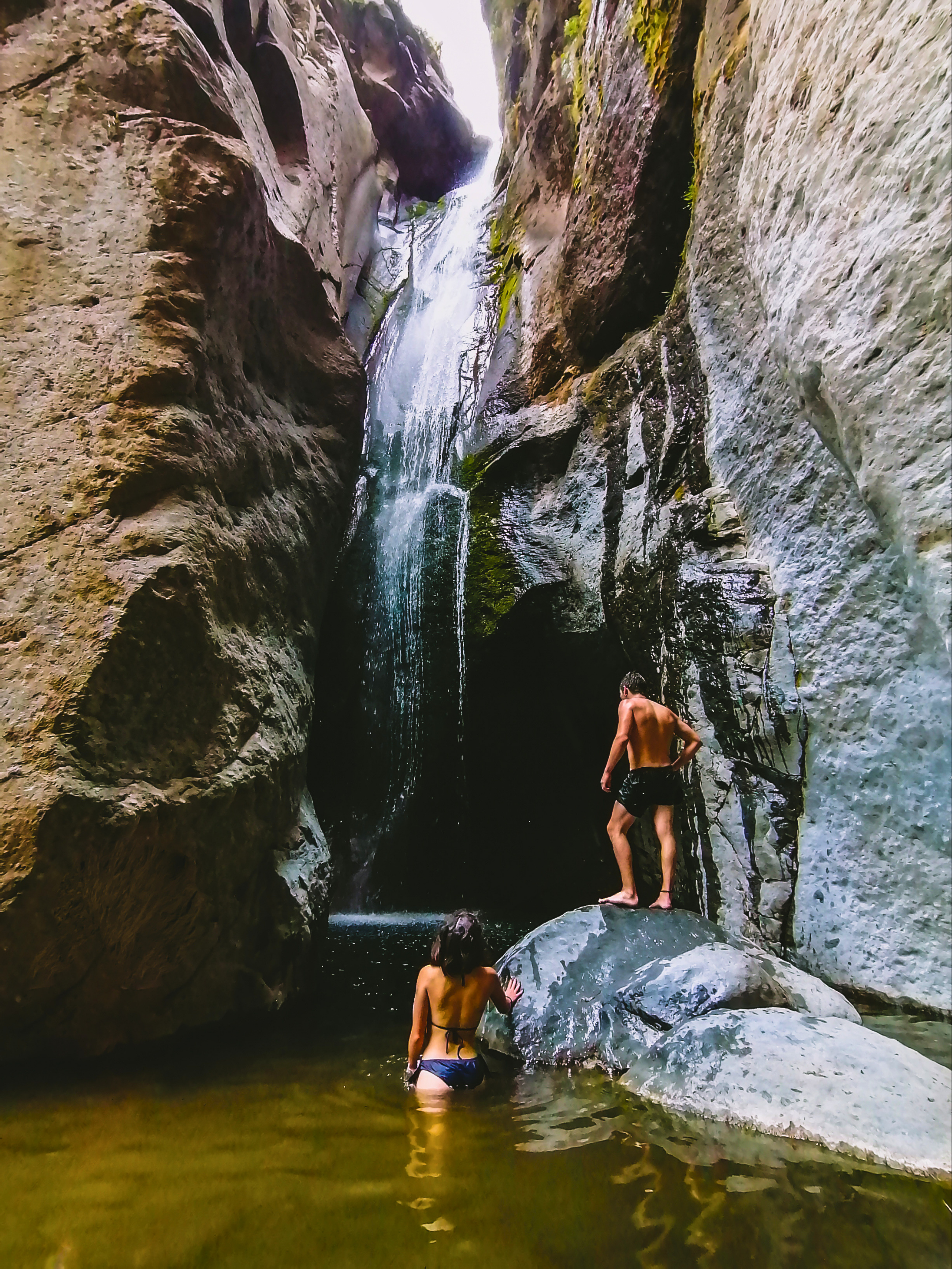 Personas disfrutando de un cañón con agua y una pequeña cascada, rodeados de paredes rocosas y vegetación.