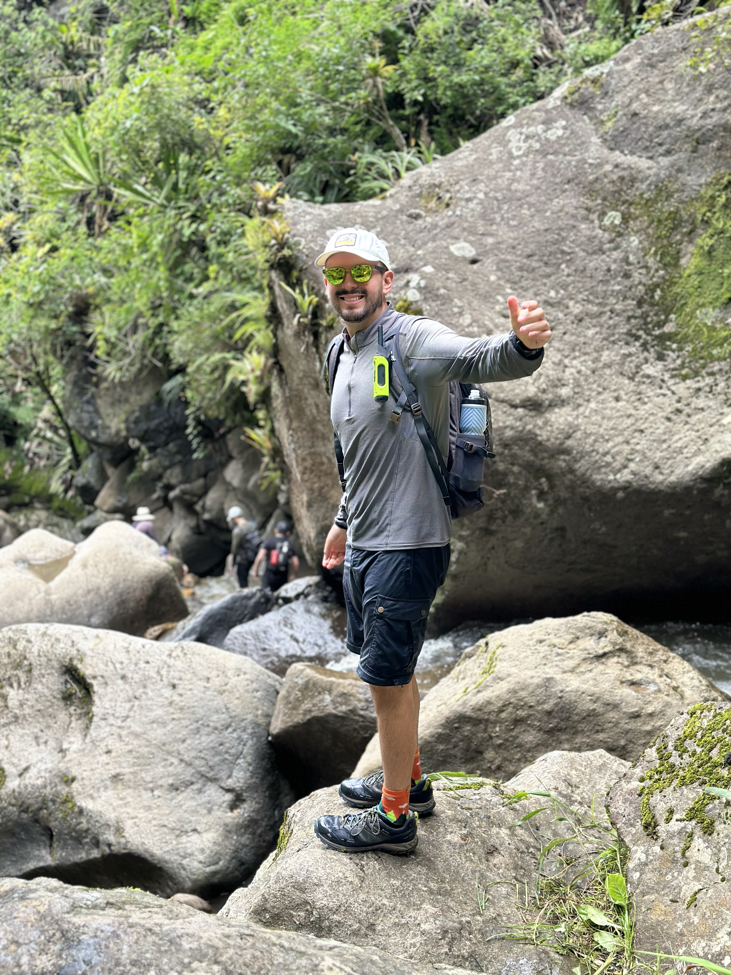 Hombre haciendo senderismo en un paisaje de rocas y naturaleza, con gorra, gafas de sol y mochila, sonriendo y mostrando un pulgar hacia arriba.
