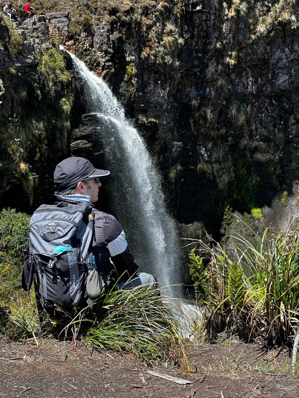 Persona con mochila y gafas mirando una cascada en un paisaje natural con vegetación.