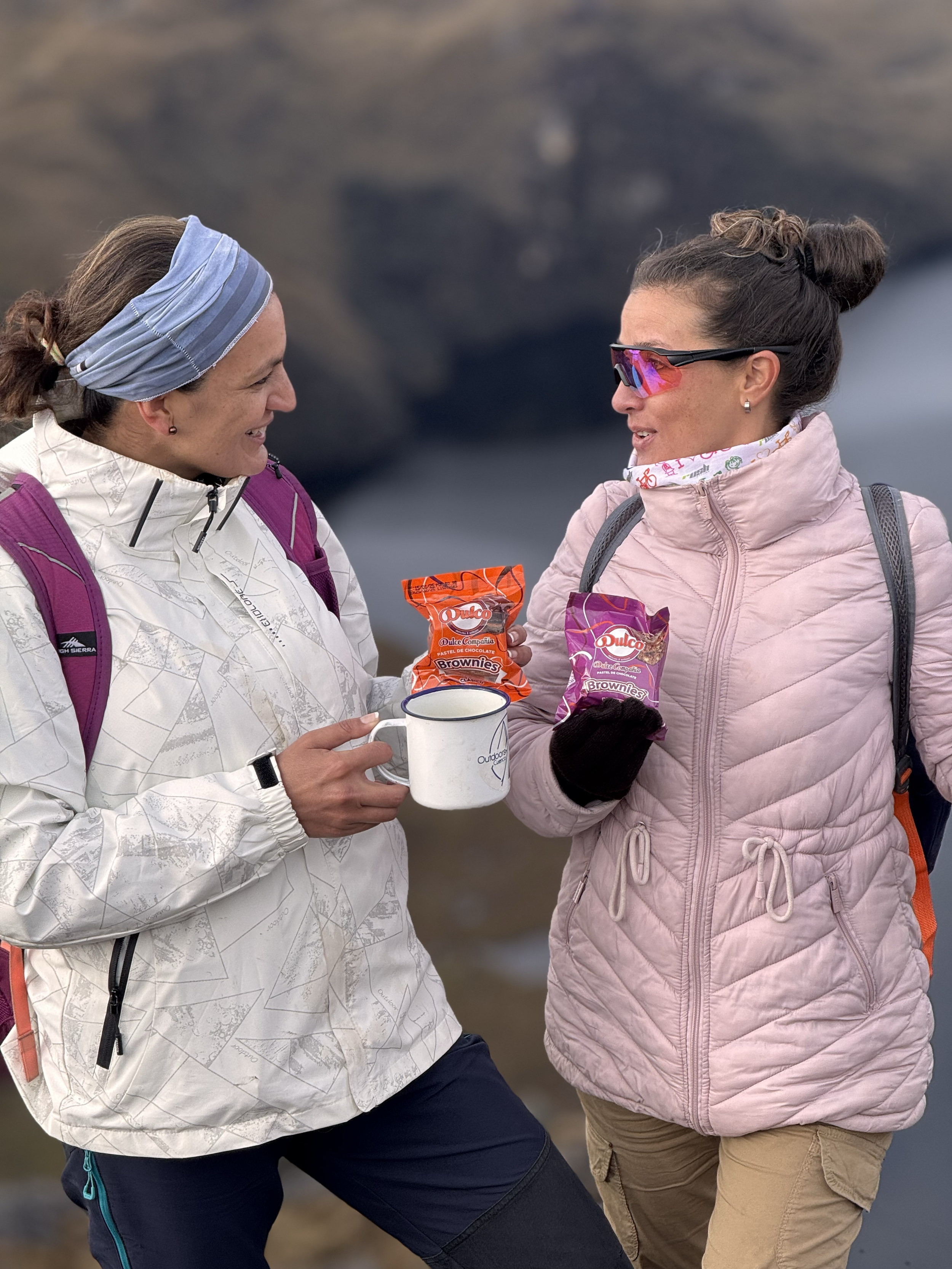 Dos mujeres en ropa de senderismo compartiendo dulces y una taza, con un paisaje de naturaleza de fondo.