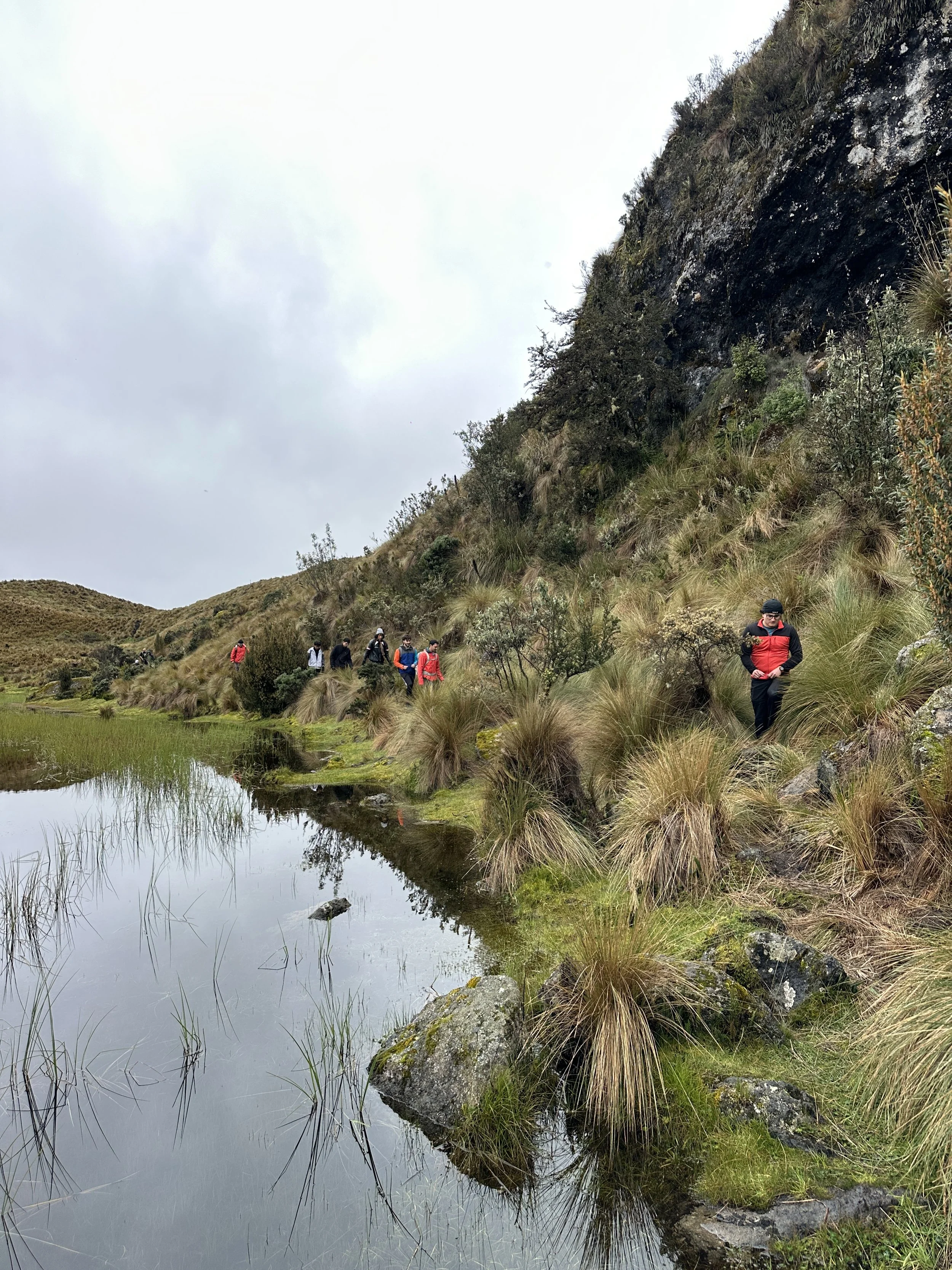 Grupo de personas caminando por un sendero a lo largo de un cuerpo de agua en un paisaje montañoso con vegetación y un cielo nublado.
