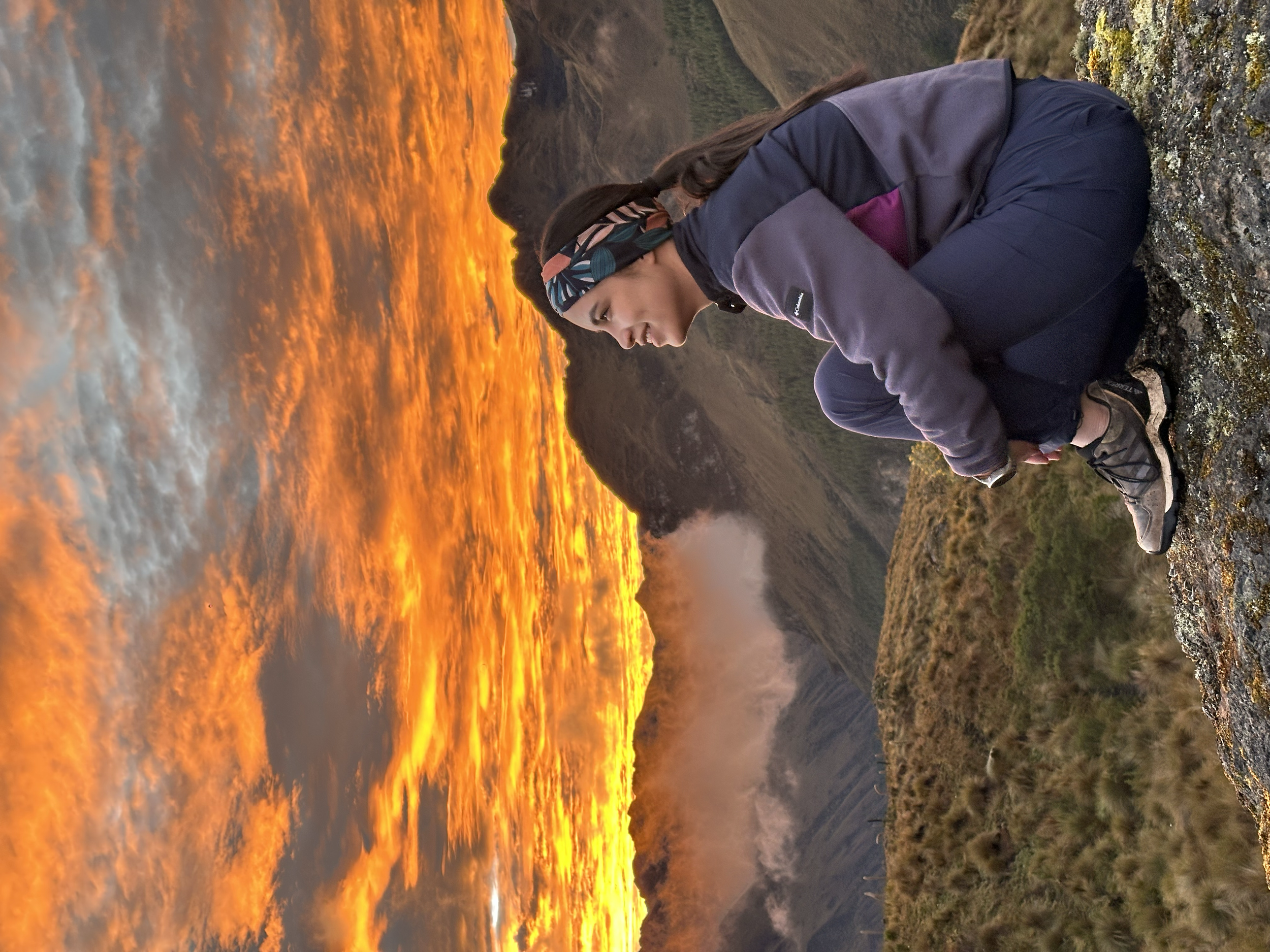 Una mujer en ropa de senderismo en cuclillas en una cima de montaña con un fondo de cielo al atardecer con nubes anaranjadas y montañas.