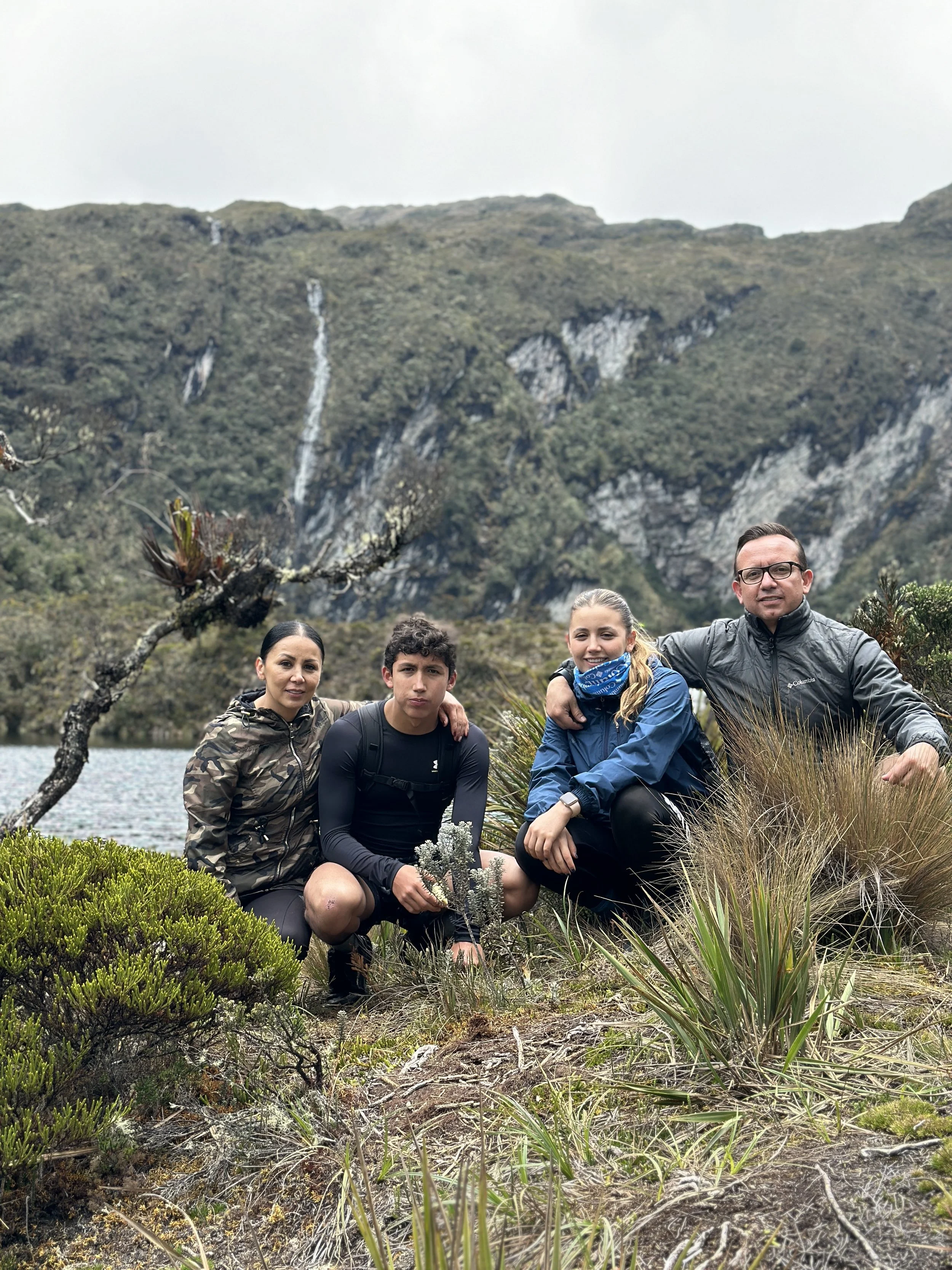 Cuatro personas en un paisaje montañoso junto a un lago, rodeadas de vegetación, durante una caminata o excursión al aire libre.