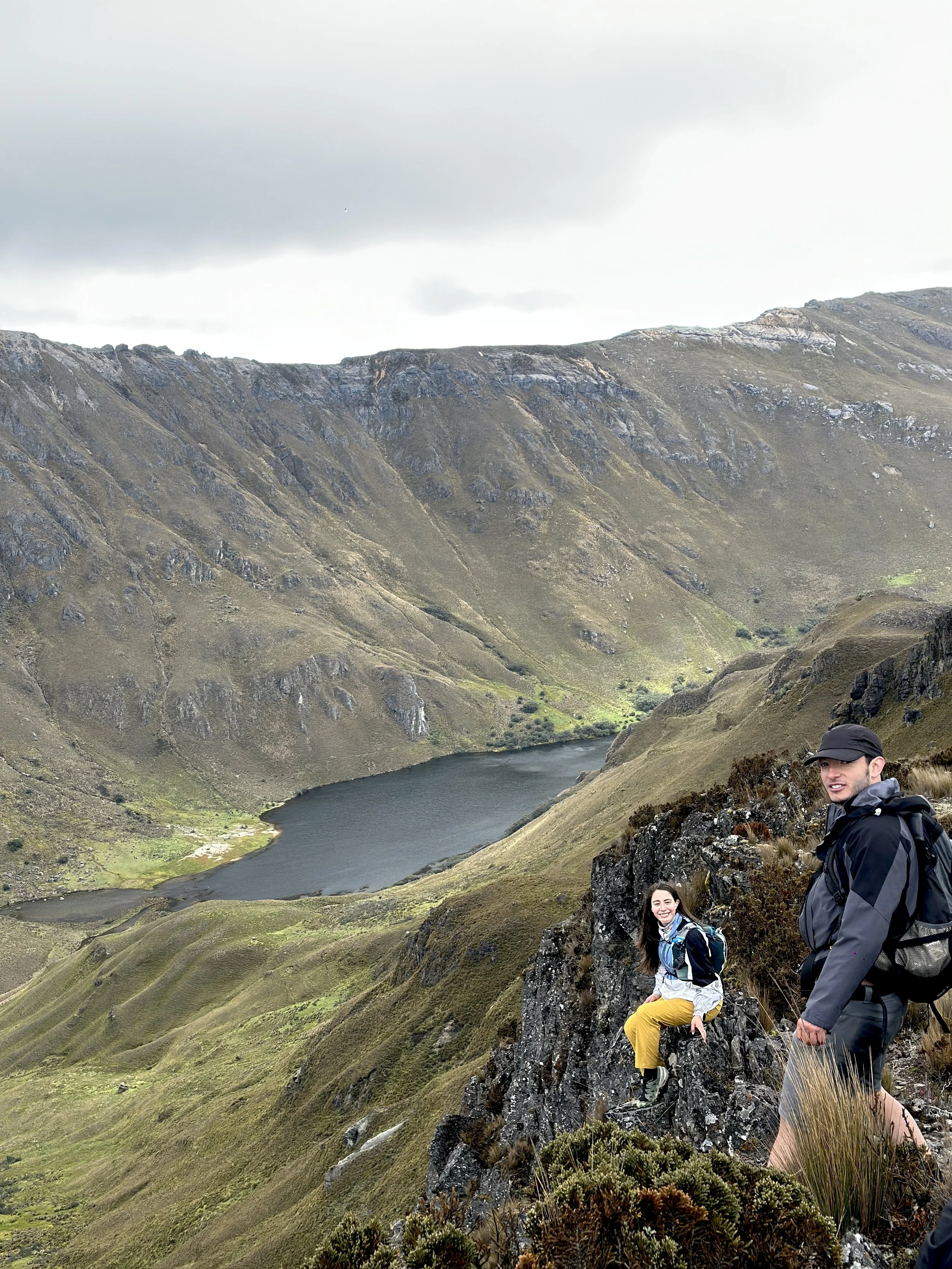 Dos excursionistas en una montaña con vista a un lago rodeado de colinas y picos rocosos, bajo un cielo nublado.