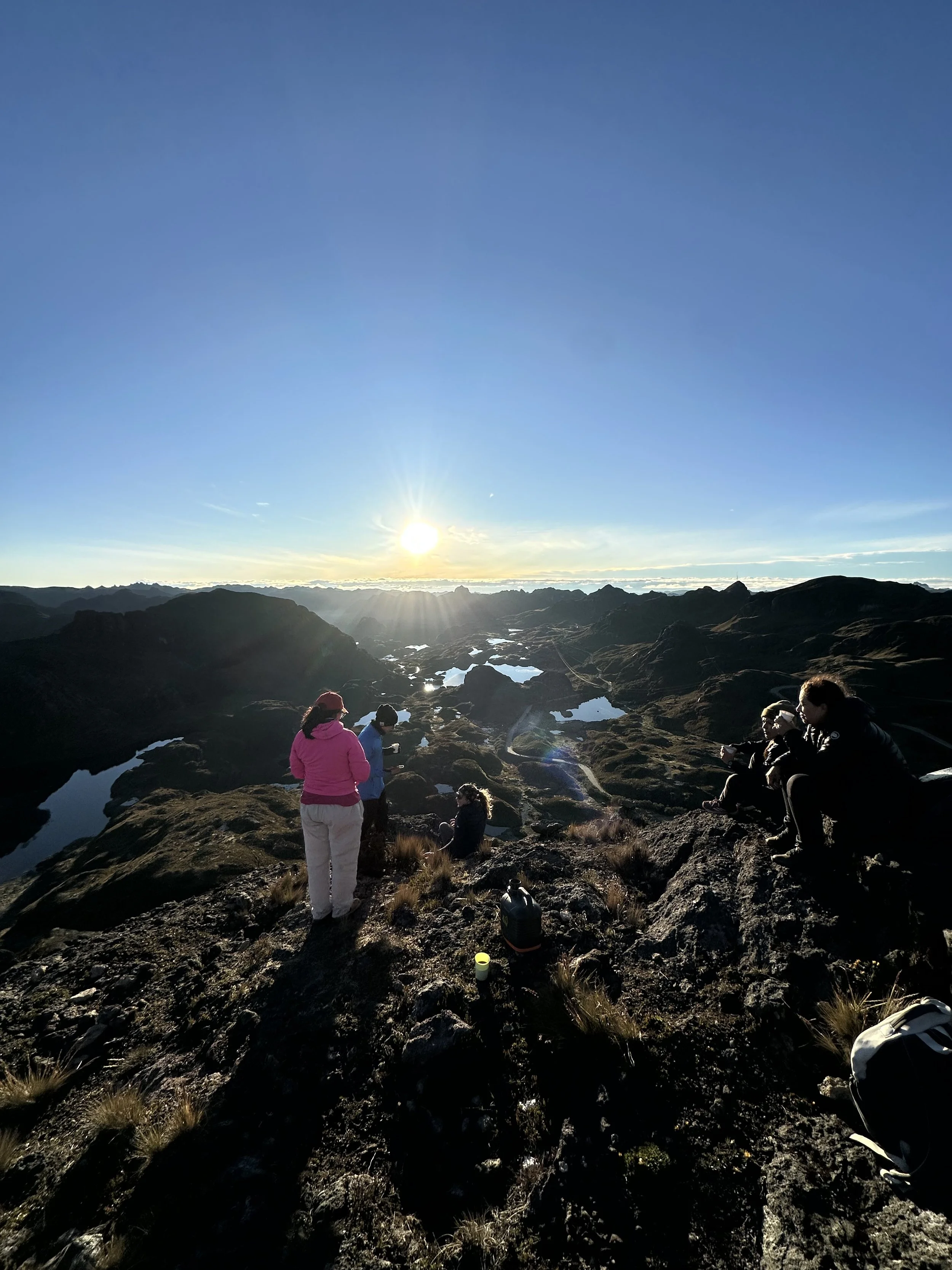 Grupo de personas en un paisaje montañoso en atardecer, algunas sentadas y otras de pie, disfrutando de la vista y tomando fotos, con charcos de agua en el suelo y montañas al fondo.