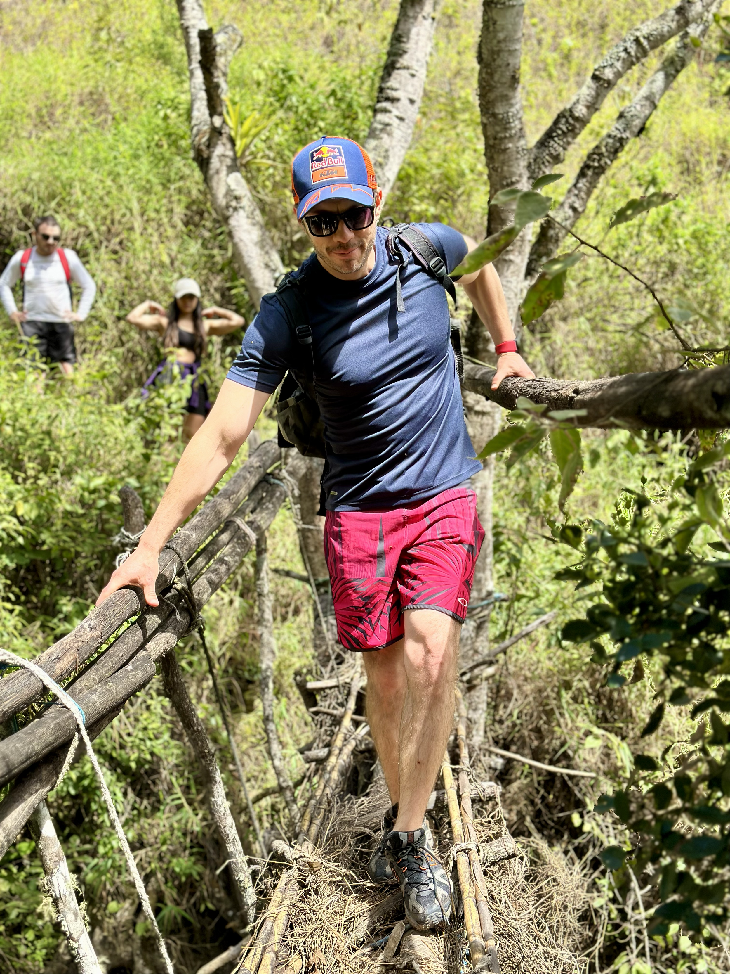 Hombre cruzando un puente de troncos en medio de un bosque, acompañado por dos personas más al fondo.