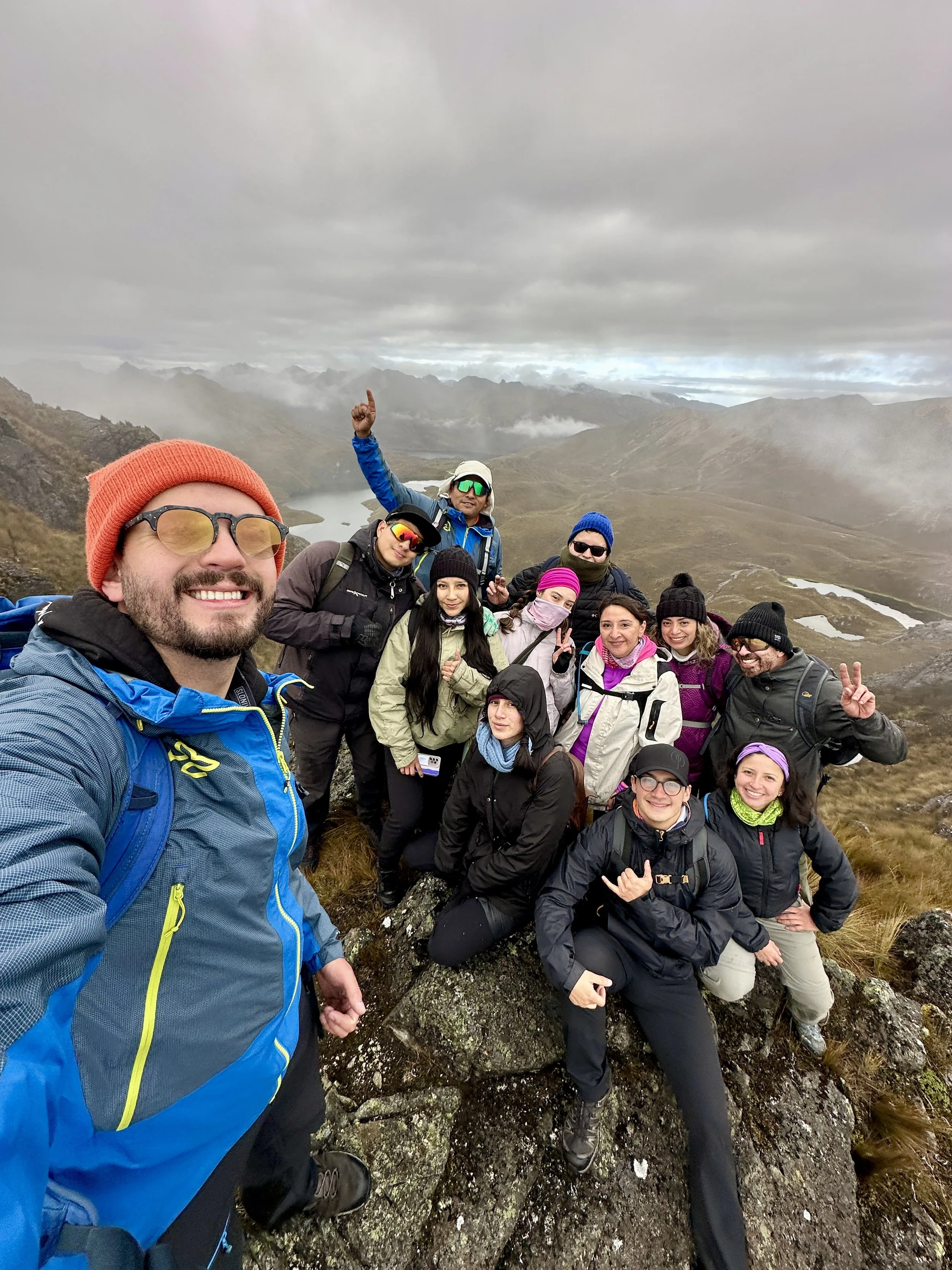 Grupo de personas haciendo senderismo en una cima de montaña con vista a lagos y picos nevados a lo lejos, en clima nublado.