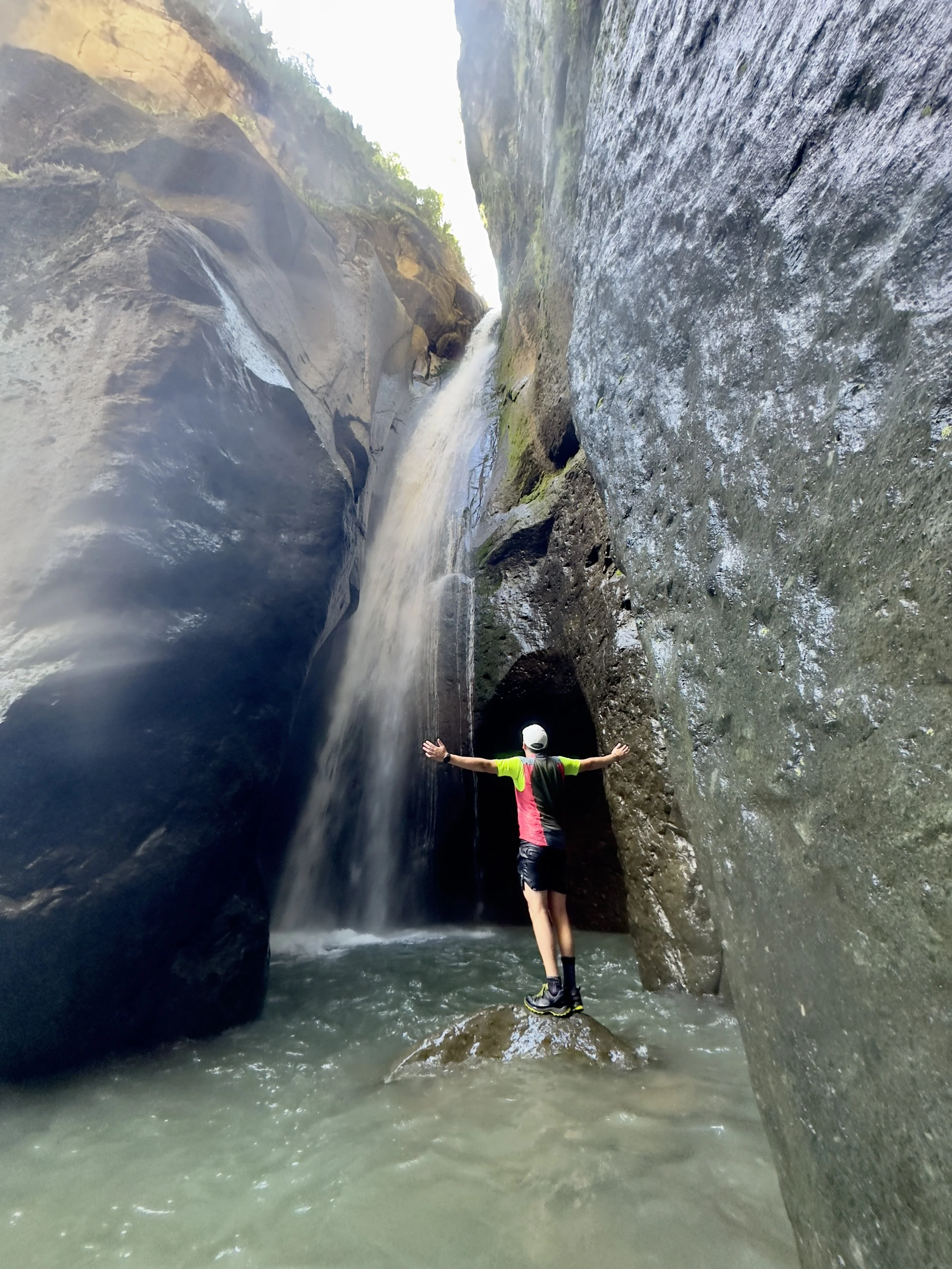 Persona de espaldas con brazos abiertos, vistiendo camiseta de manga larga de color rosa y negro, pantalones cortos y casquito, de pie en una roca en medio de una poza de agua, rodeada de paredes de roca y un pequeño salto de agua en un cañón.