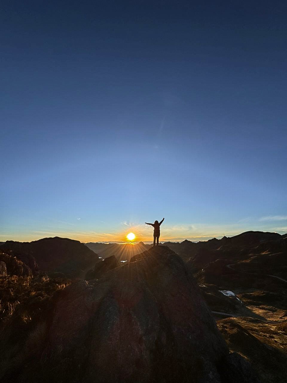 Persona con brazos extendidos en cima de una roca en un paisaje de montañas durante el amanecer o atardecer, cielo despejado con el sol cerca del horizonte.