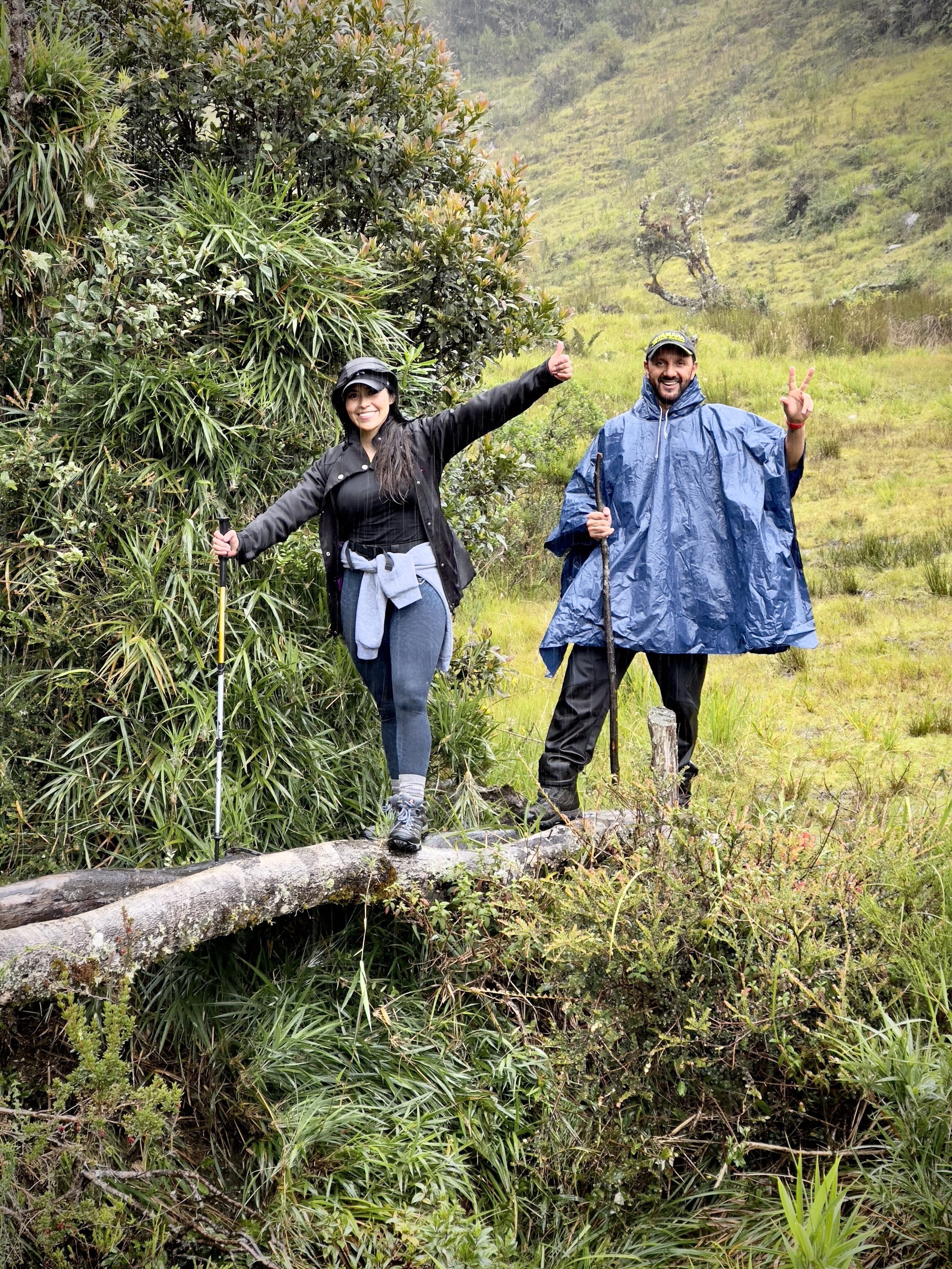 Dos personas sonrientes en ropa de lluvia, haciendo senderismo en un entorno natural con vegetación abundante y colinas en el fondo.