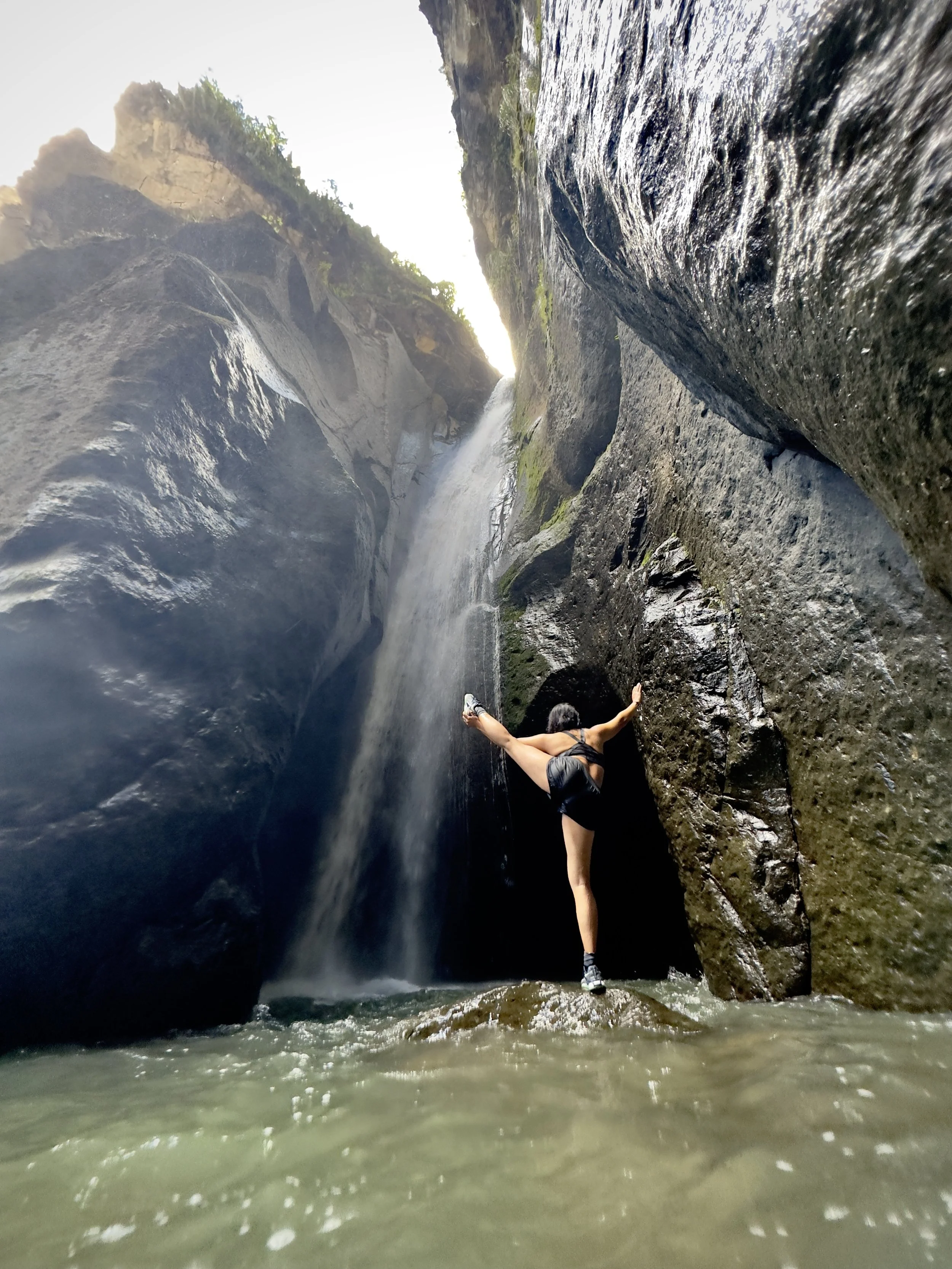 Persona haciendo footing en un cañón con paredes empinadas y agua en el suelo, con una cascada en la parte superior, vista desde el nivel del agua.