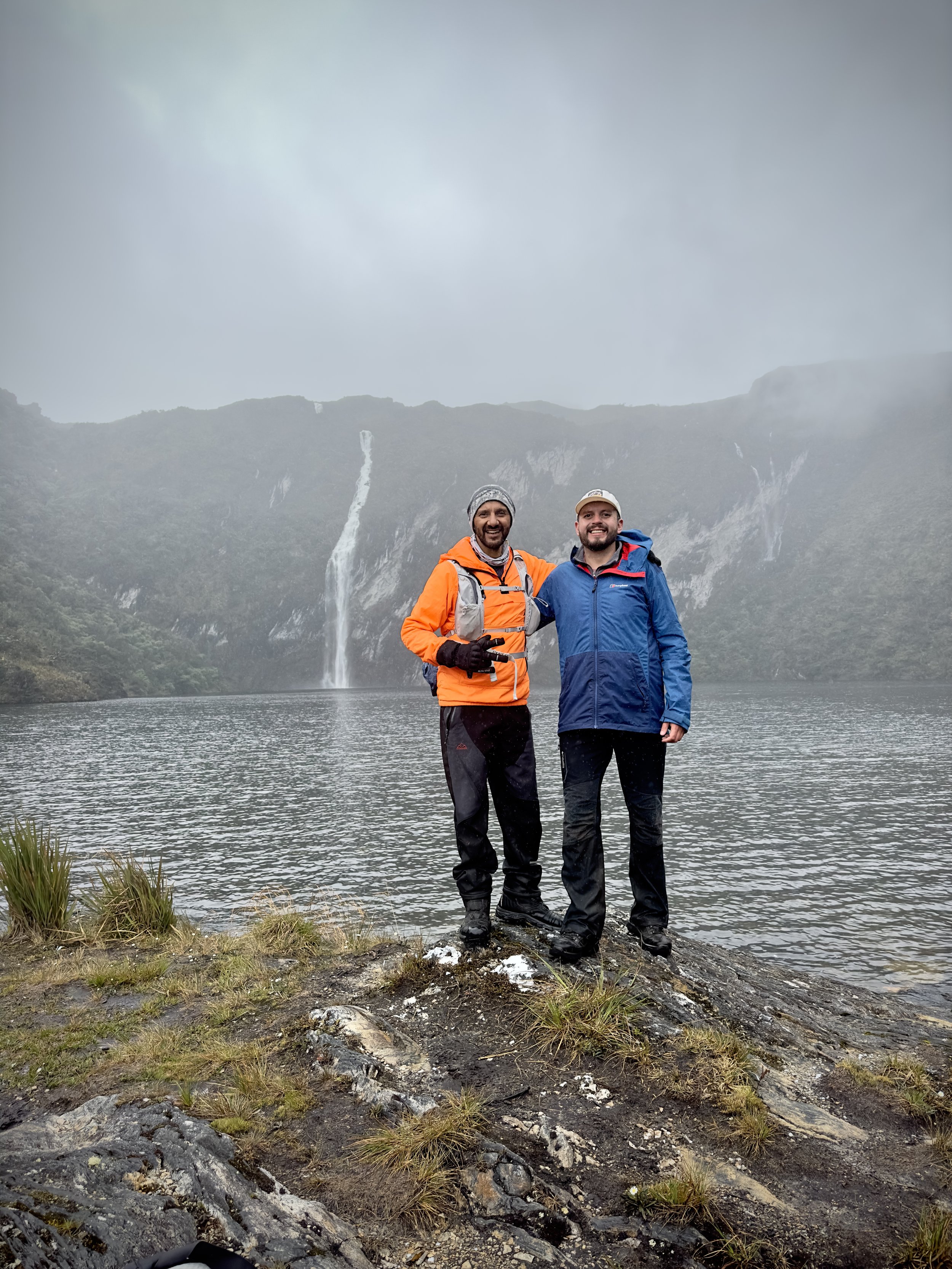 Dos hombres con ropa de invierno y mochilas en un paisaje de lago y cascada en un entorno montañoso