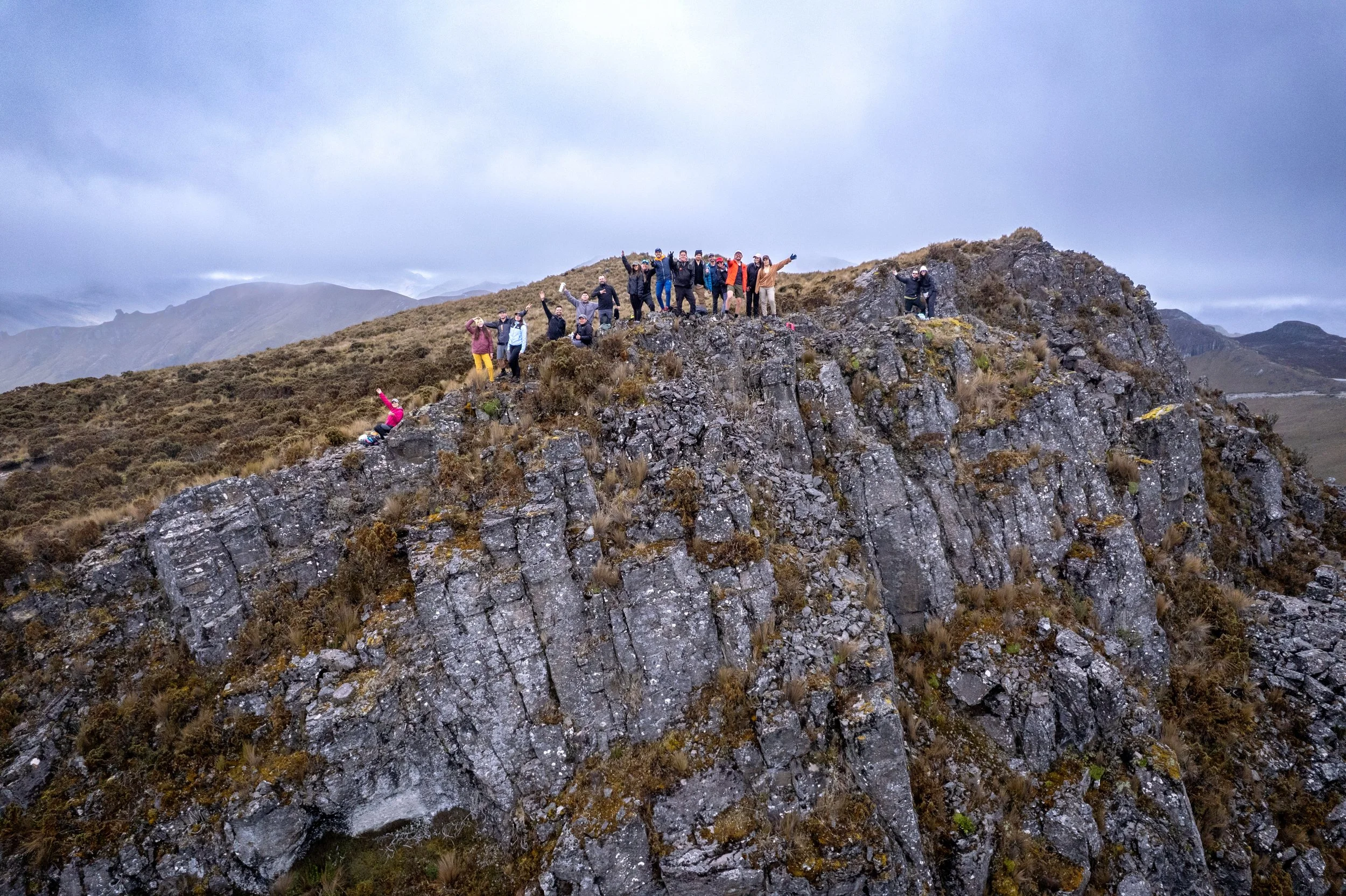 Grupo de personas en la cima de una montaña rocosa con un paisaje montañoso en el fondo, algunos toman fotos y otros celebran.