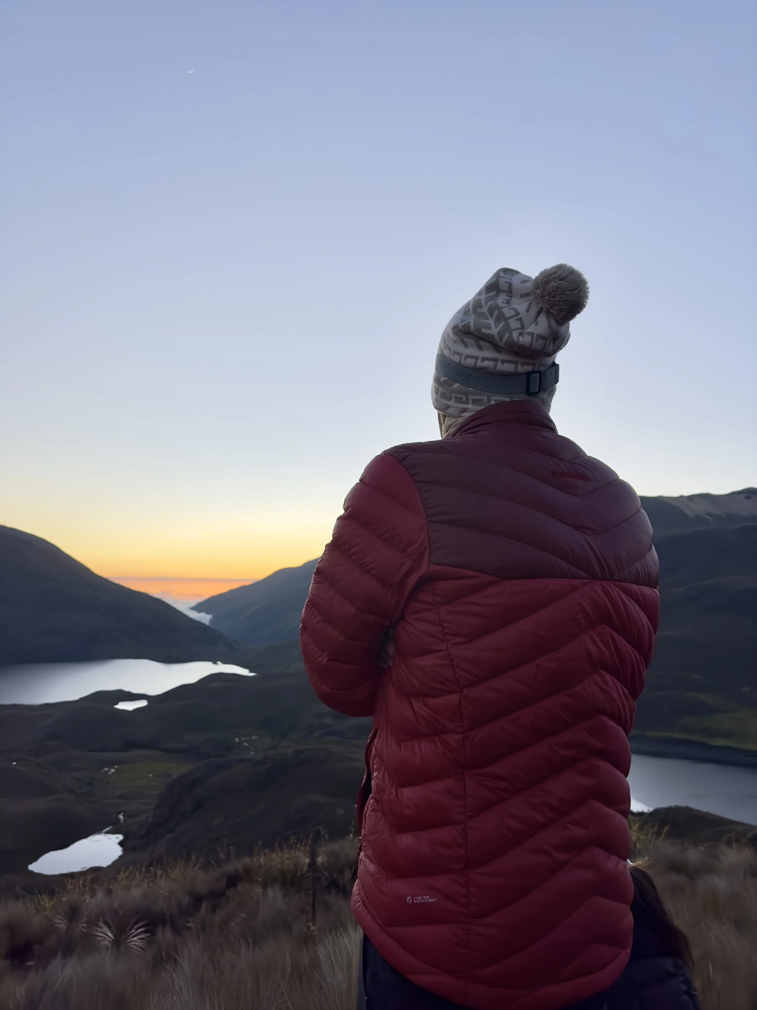 Persona con abrigo rojo y gorro mirando hacia el atardecer en un paisaje montañoso con lagos.