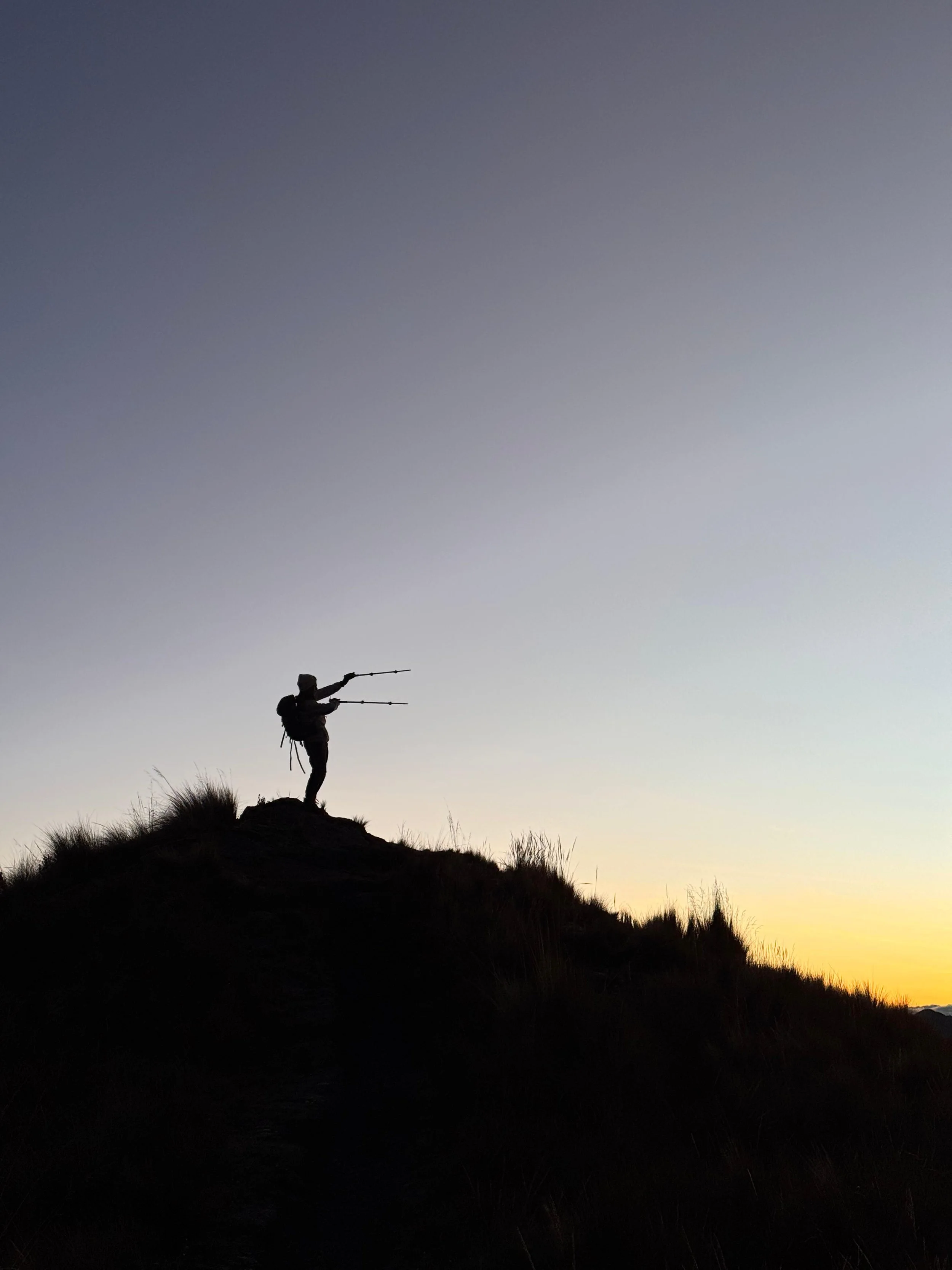 Silhouette de una persona con mochila en la cima de una colina, apuntando con dos bastones hacia el horizonte al atardecer.