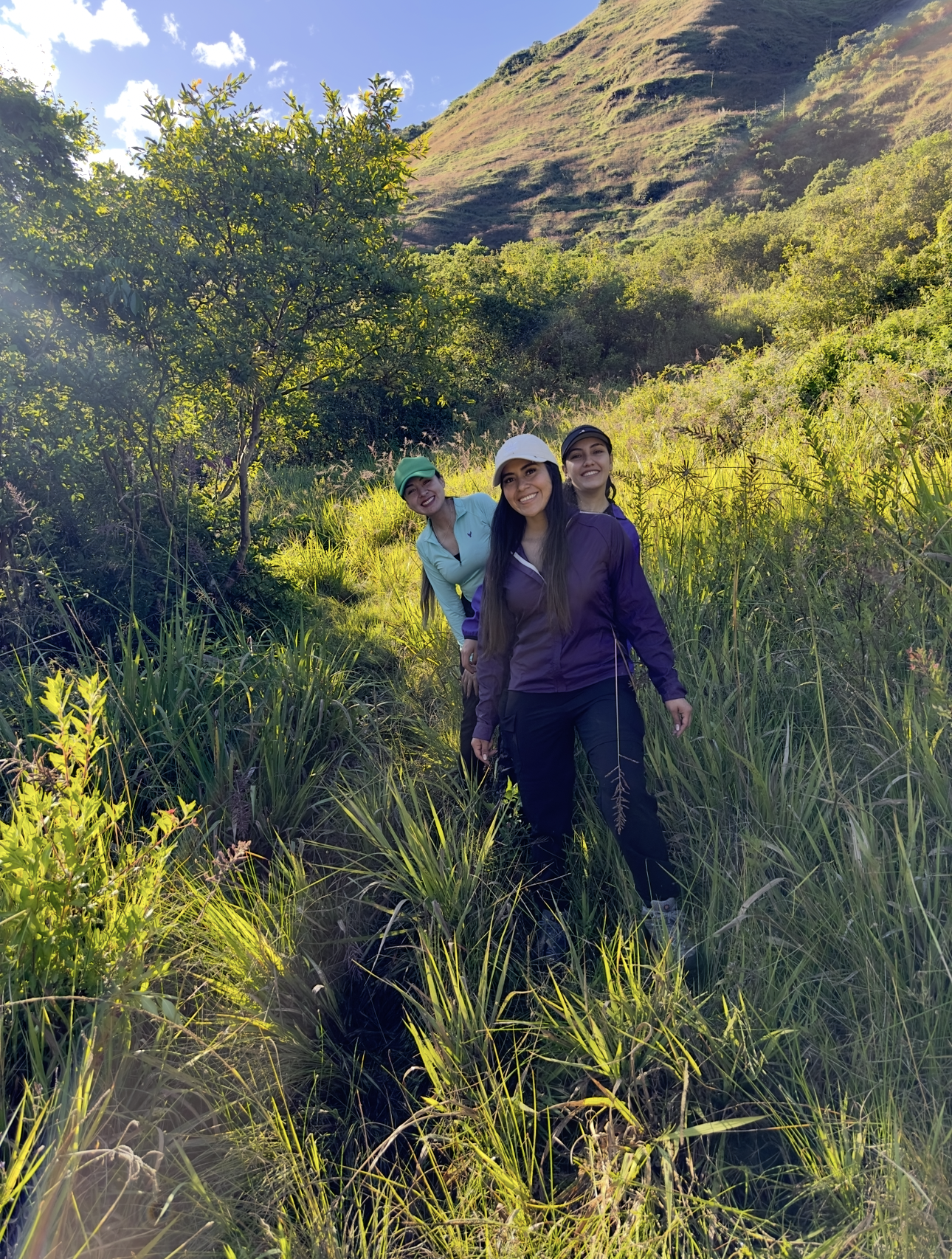 Tres mujeres caminando por un sendero en un entorno natural con vegetación y una montaña al fondo, en un día soleado.