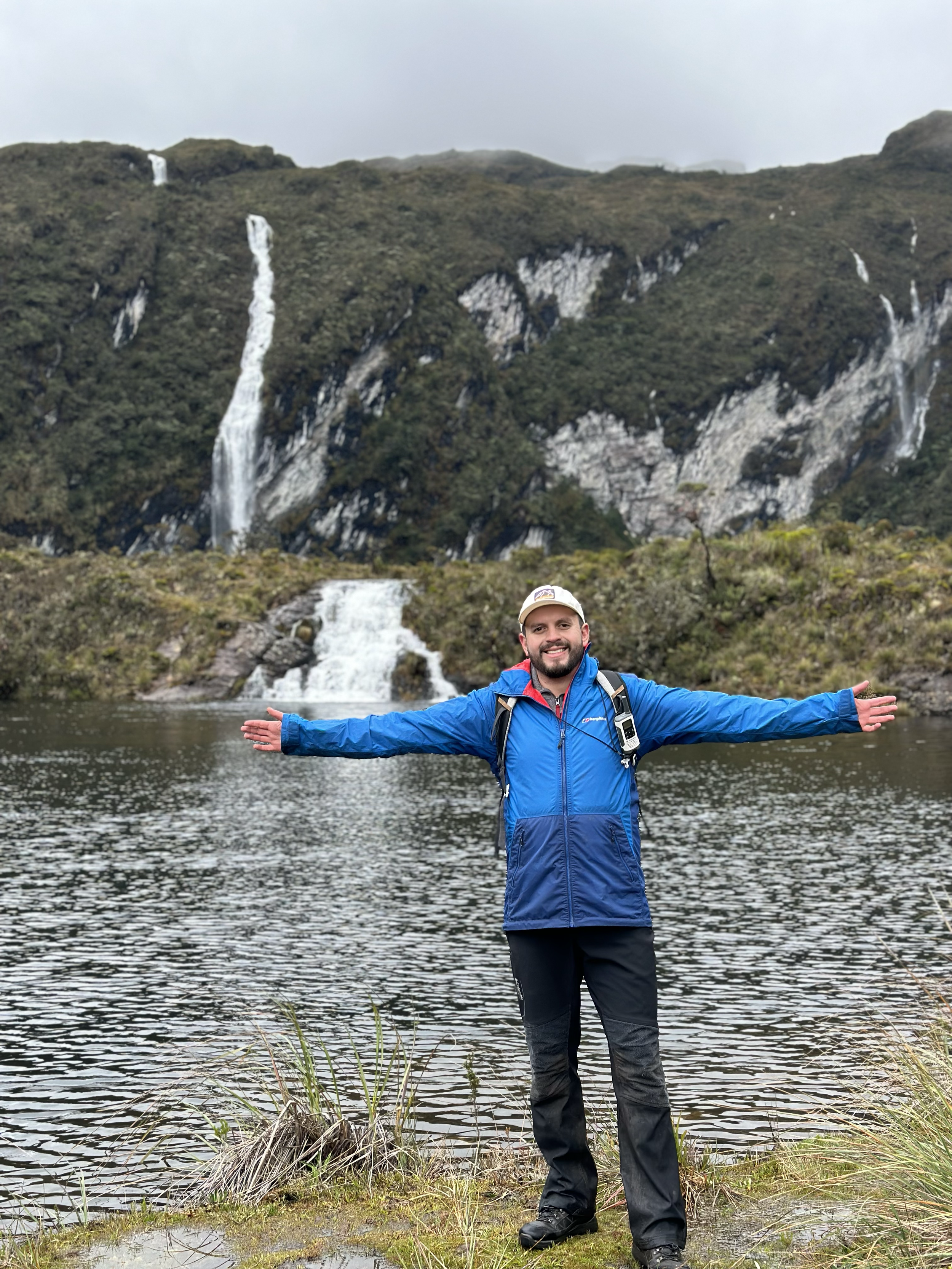 Hombre sonriente con chaqueta azul y mochila en un lago con cascadas de fondo en un paisaje natural.