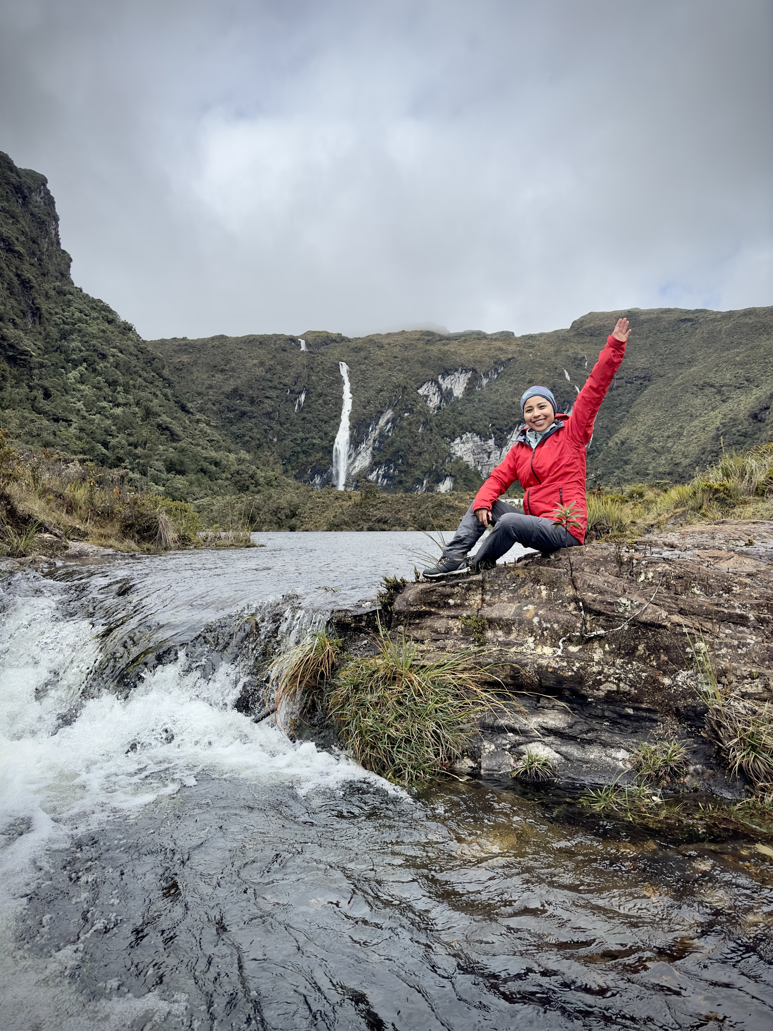 Mujer en ropa de senderismo en un río de montaña con cascadas al fondo, sonriendo y saludando con una mano.