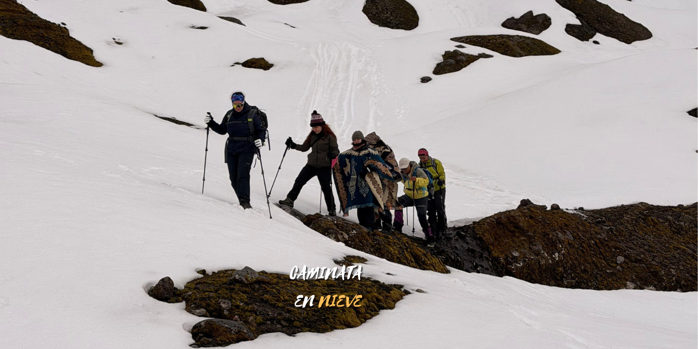 Grupo de personas haciendo caminata en la nieve en un paisaje de montaña con rocas. Algunas personas llevan bolsas y ropa de abrigo, y usan bastones de senderismo.