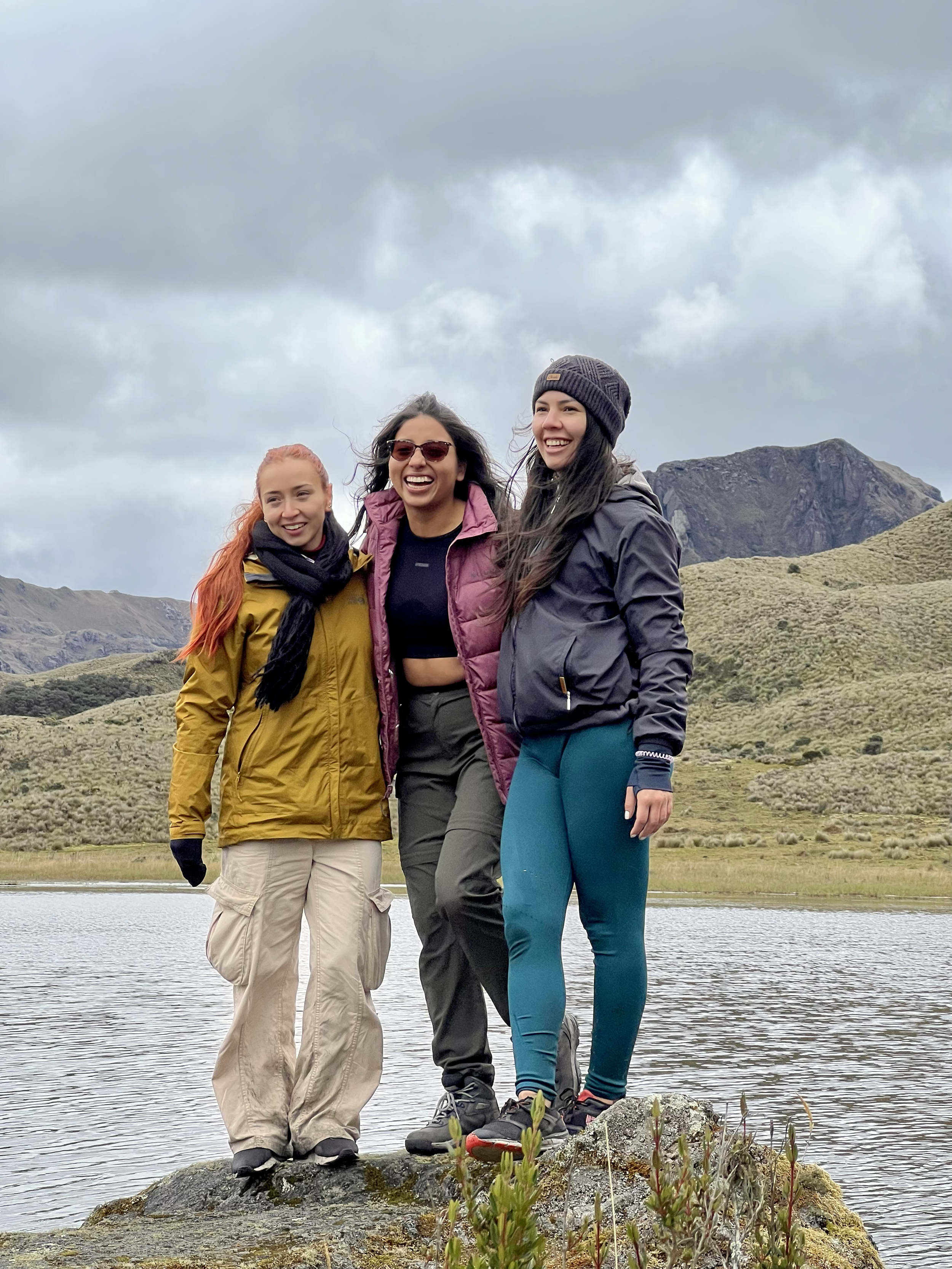 Tres mujeres sonriendo y posando juntas en un entorno natural con agua y montañas, vestidas con ropa de abrigo para senderismo.
