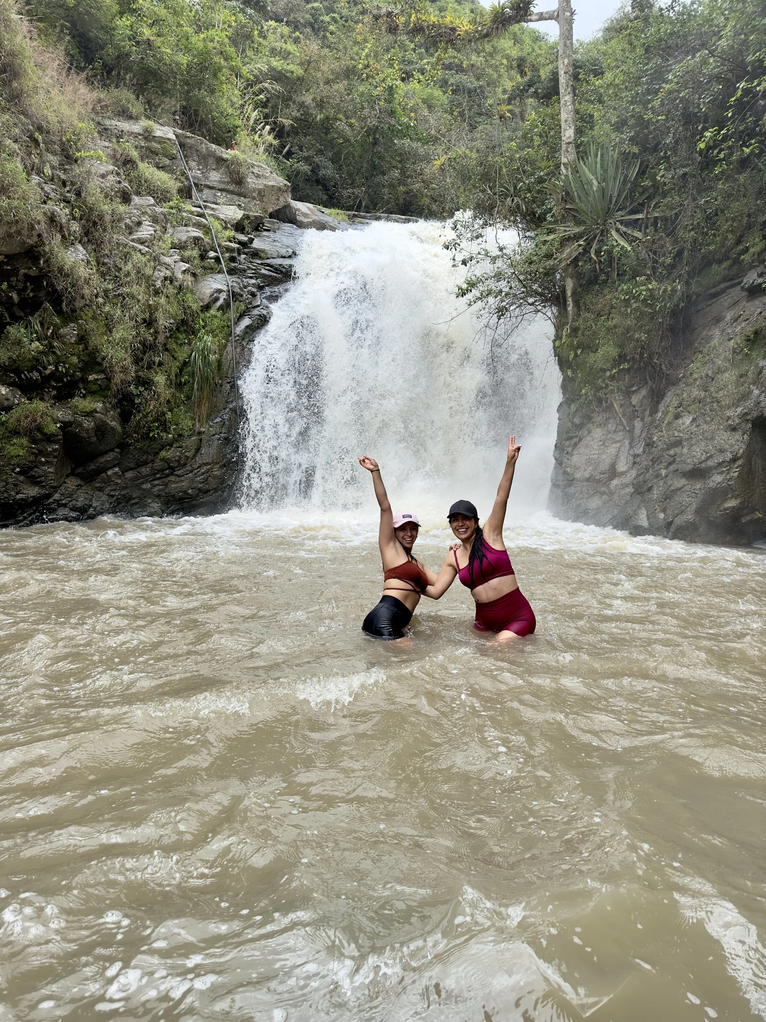Dos mujeres disfrutando en un río con una cascada en el fondo, rodeadas de vegetación verde.