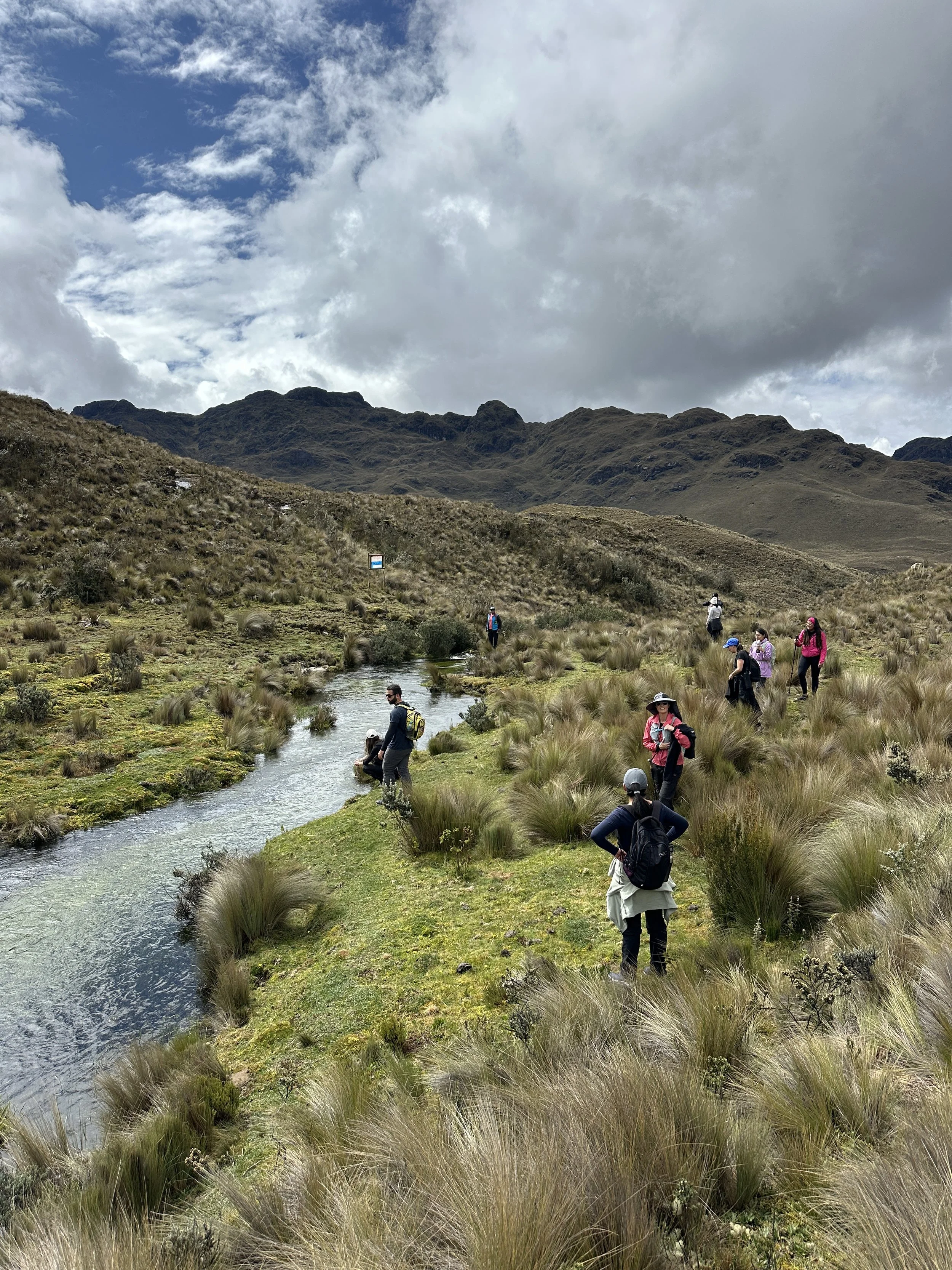 Grupo de personas haciendo senderismo y observando un río en un paisaje montañoso con nubes en el cielo.