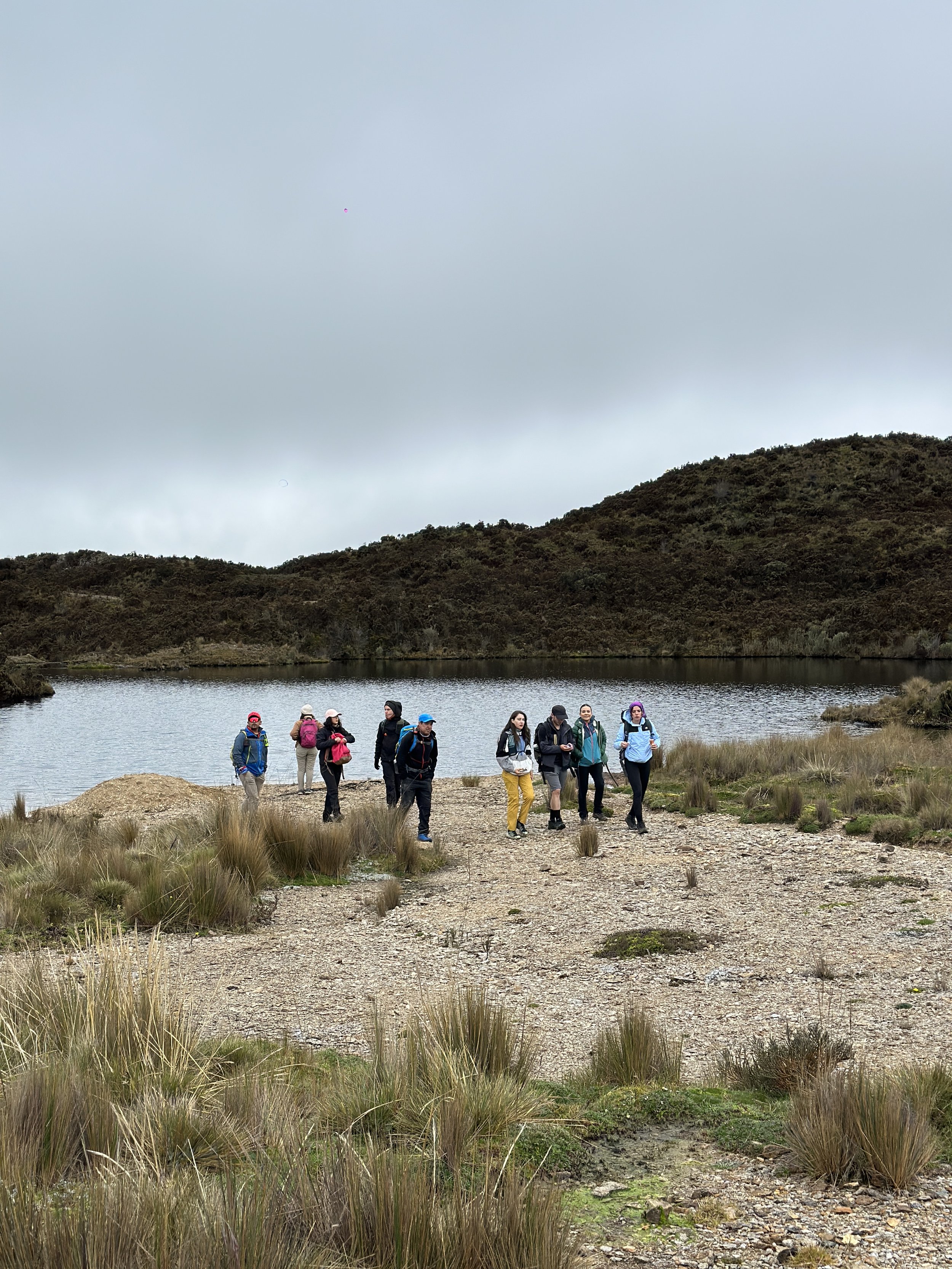 Grupo de personas haciendo senderismo cerca de un lago en un paisaje natural, con colinas y cielo nublado.