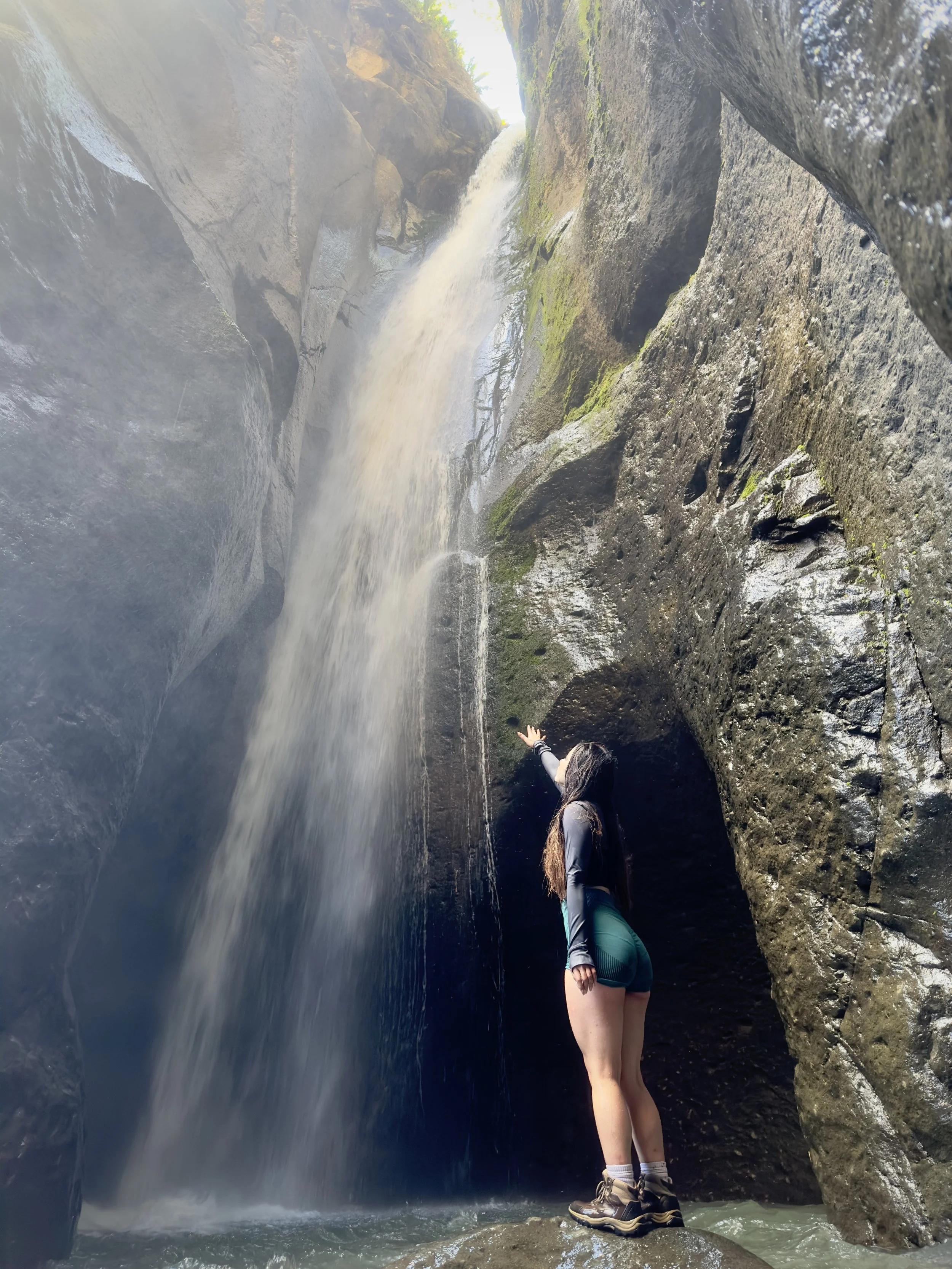 Persona de espaldas en botas de trekking, de pie junto a una roca en medio de una cascada en un cañón rocoso con agua cayendo y formando niebla.