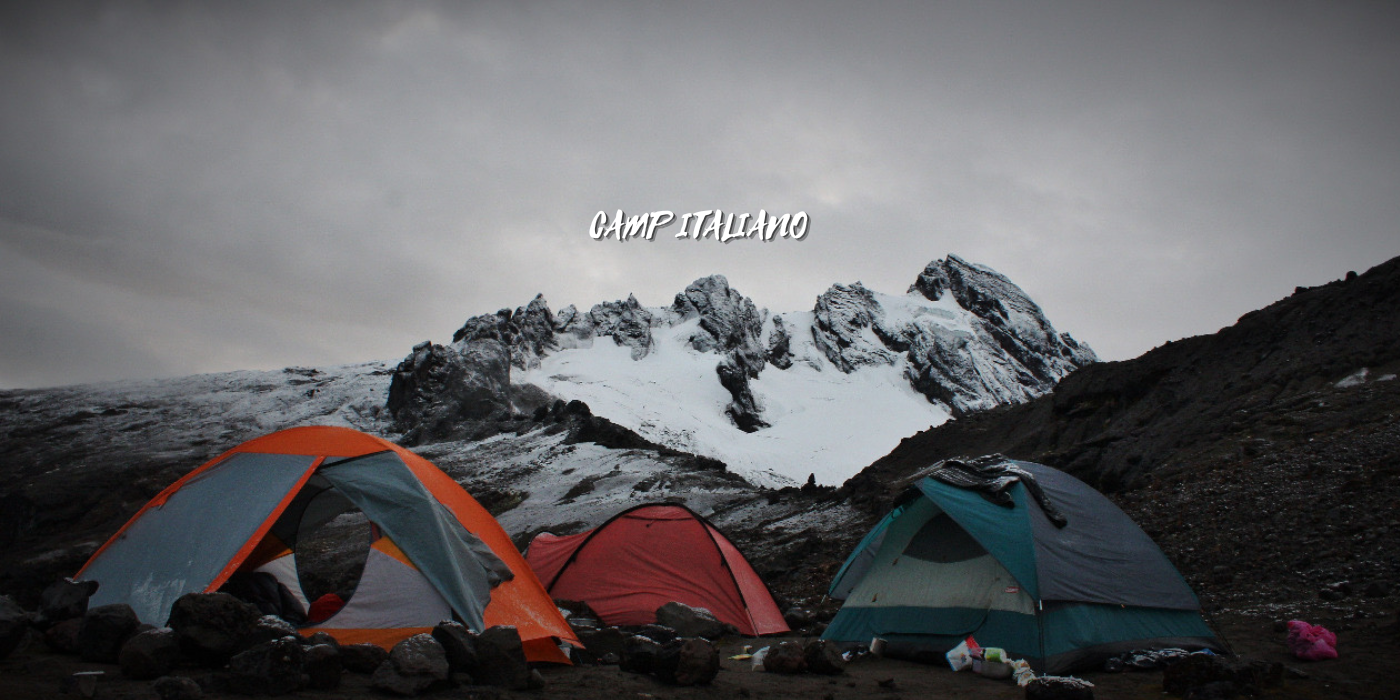 Campamento de tiendas de campaña en un entorno montañoso con picos cubiertos de nieve en el fondo y cielo nublado, en la región de Italia.