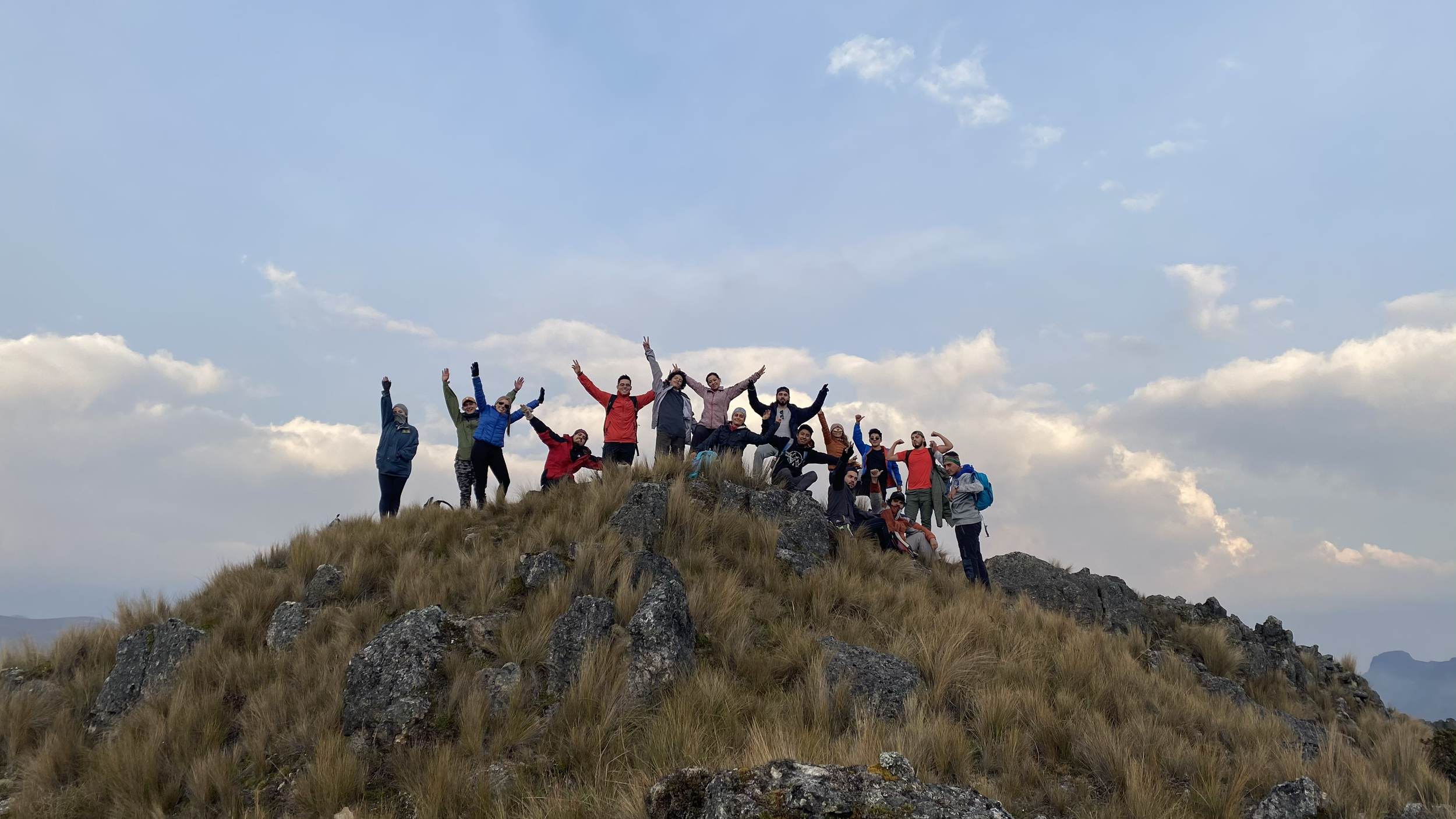Grupo de personas en la cima de una montaña, celebrando con los brazos levantados, con cielo y nubes de fondo.