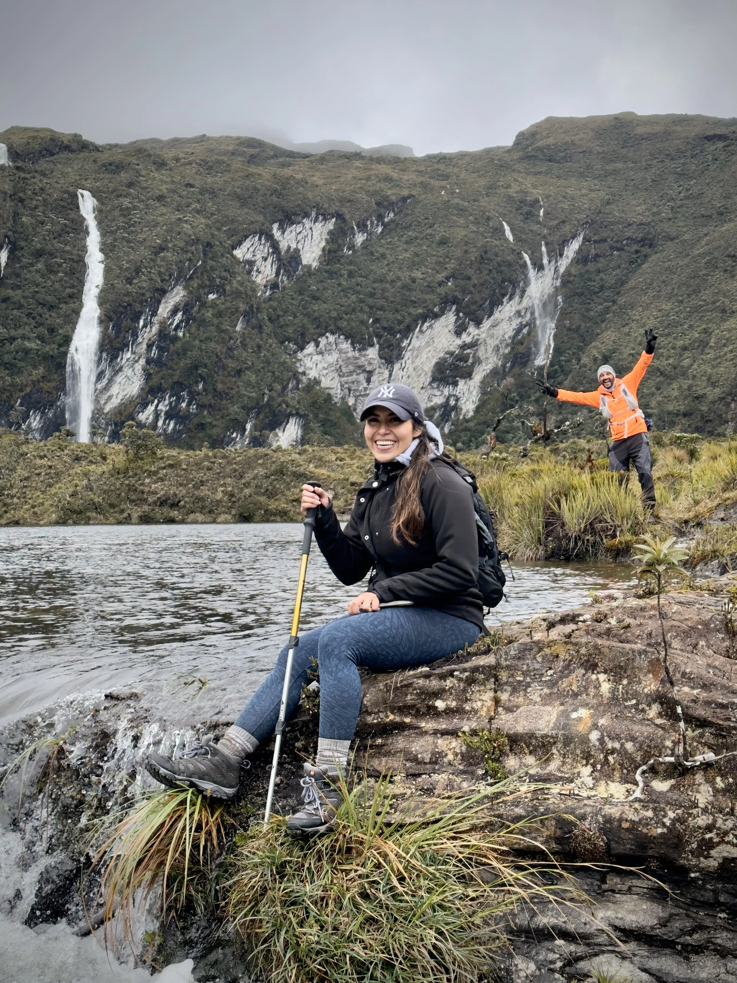 Dos personas disfrutando de un paisaje natural con cascadas en las montañas, una sentada en una roca junto a un río con una sonrisa y otra de pie con brazos extendidos y señal de victoria, ambientado en un día nublado.