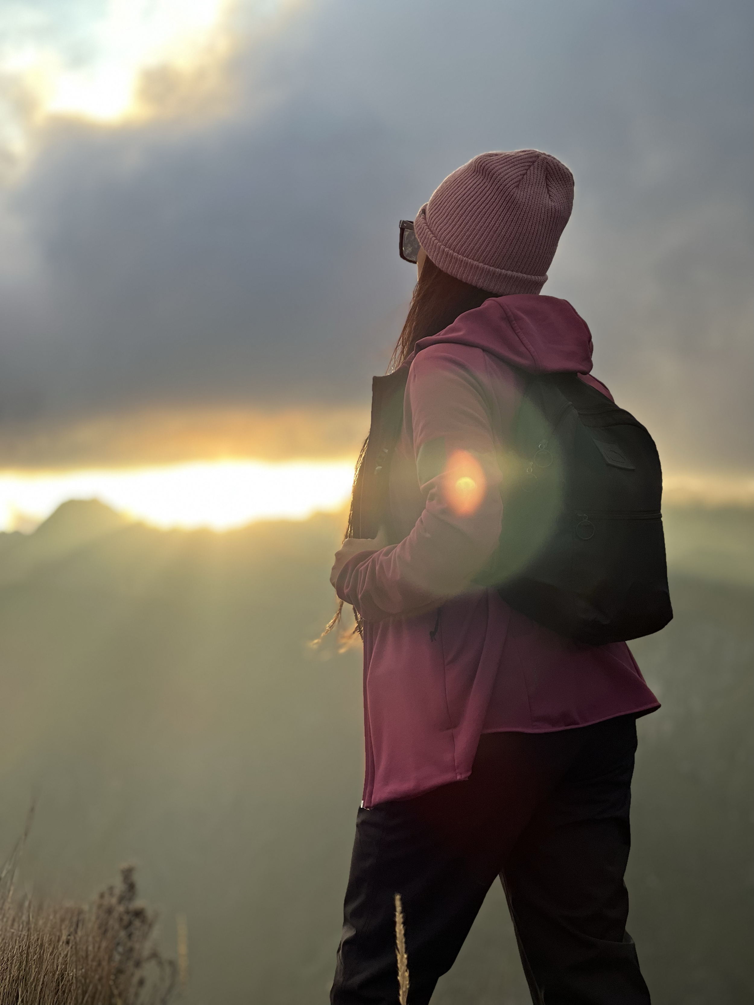 Mujer con abrigo rosa, gorro rosa y mochila negra, de espaldas, observando el atardecer en un paisaje montañoso.