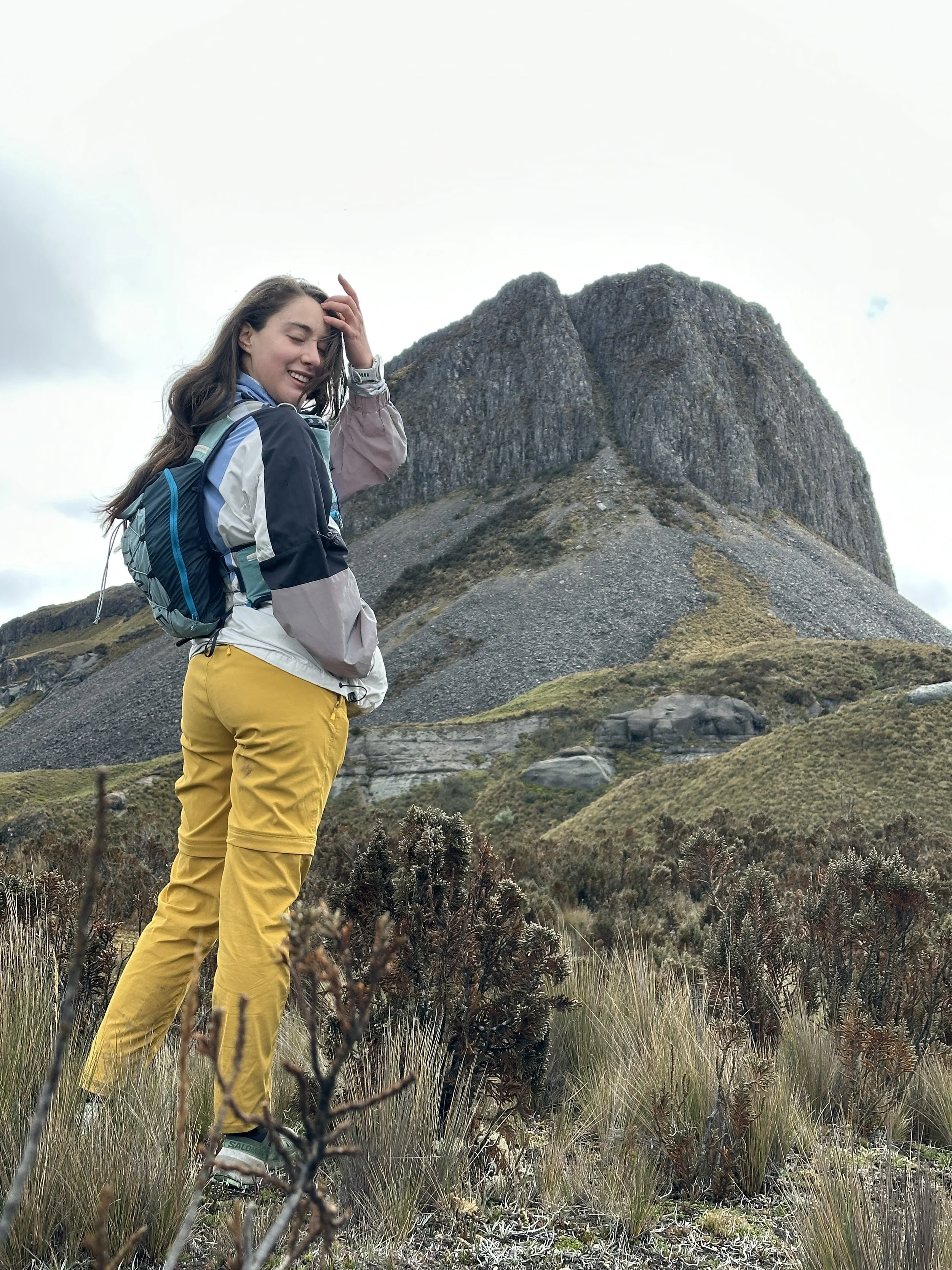 Mujer sonriendo con mochila en un paisaje de montaña y vegetación. Montañas de roca en el fondo.