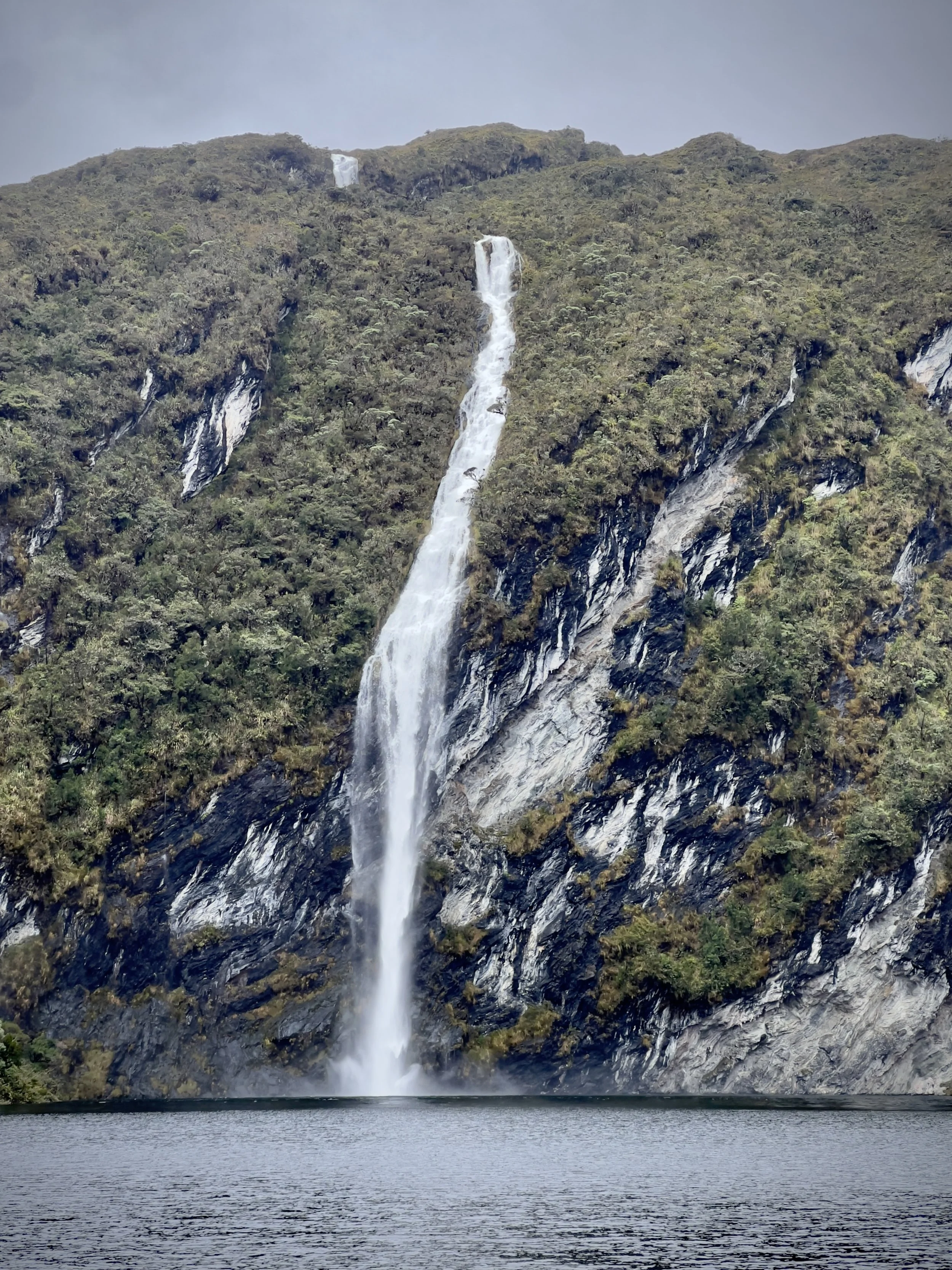 Cascada en medio de una vegetación montañosa y un lago en primer plano.