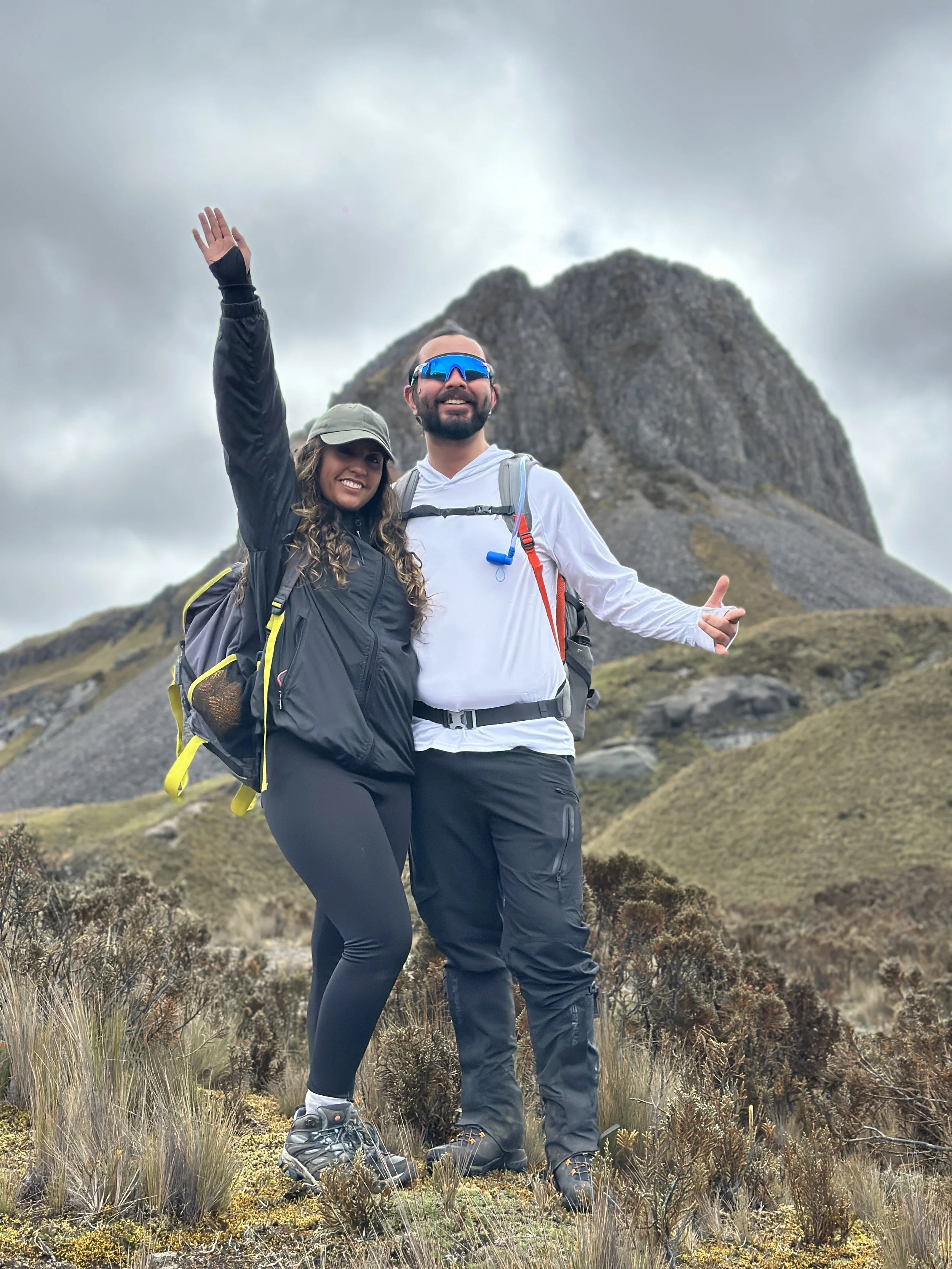 Pareja de excursionistas en la montaña, levantando las manos, con un fondo de paisaje montañoso y cielo nublado.