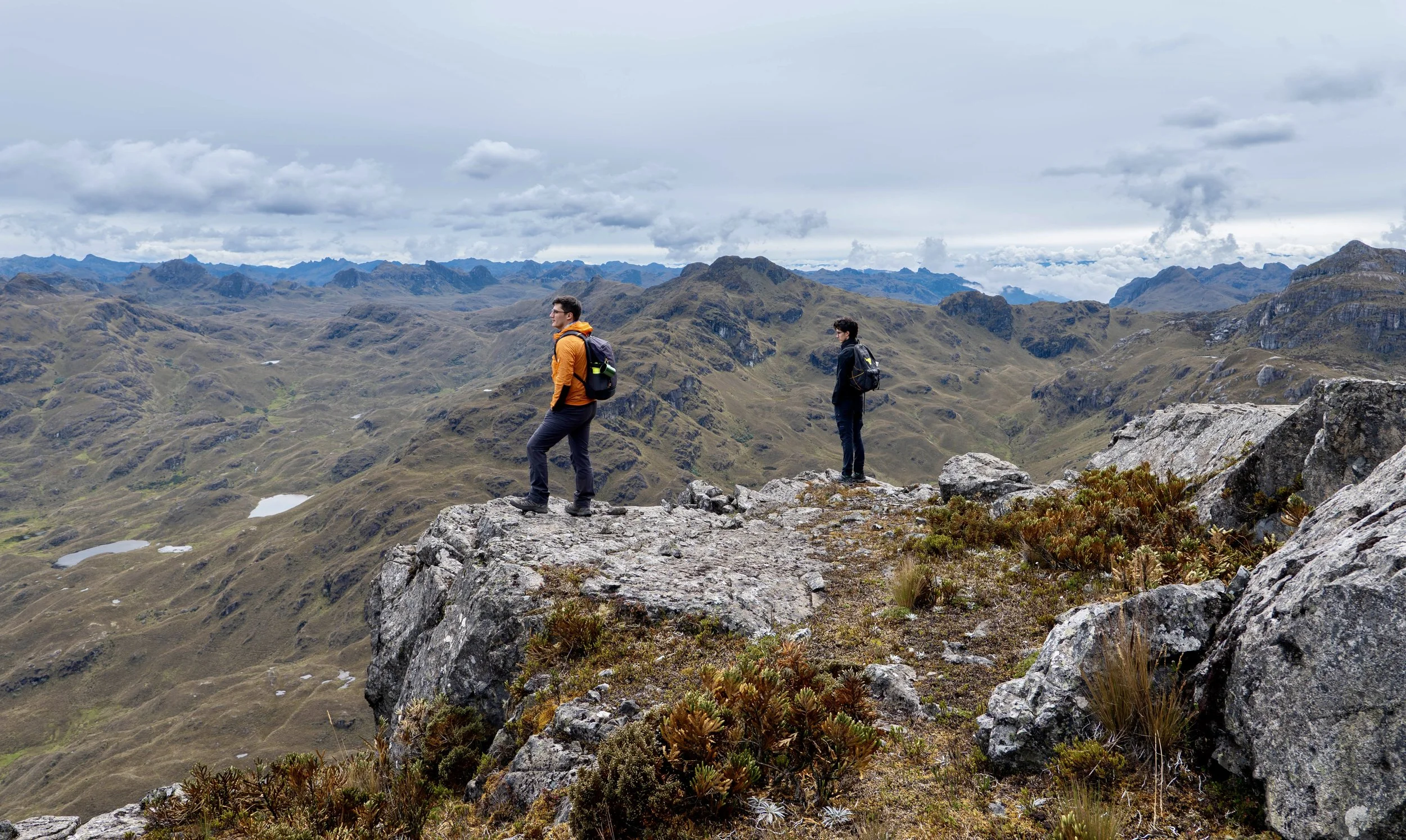 Dos personas con mochilas de senderismo en una montaña, observando el paisaje de montañas y lagunas en un día nublado.