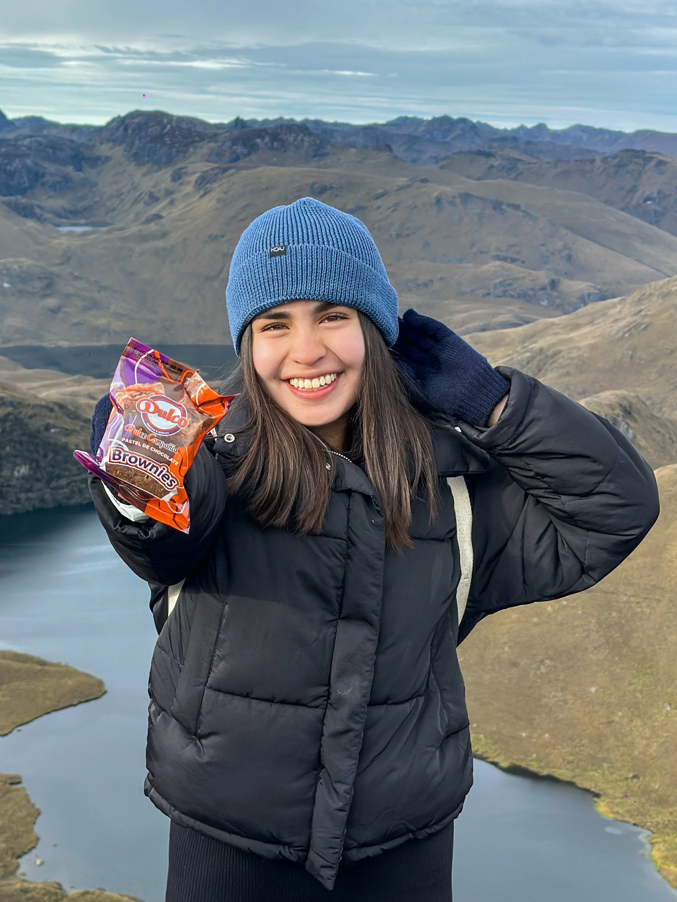Una joven sonriendo, usando un gorro azul y un abrigo negro, en un paisaje montañoso con un lago, sosteniendo una bolsa de brownies de chocolate.