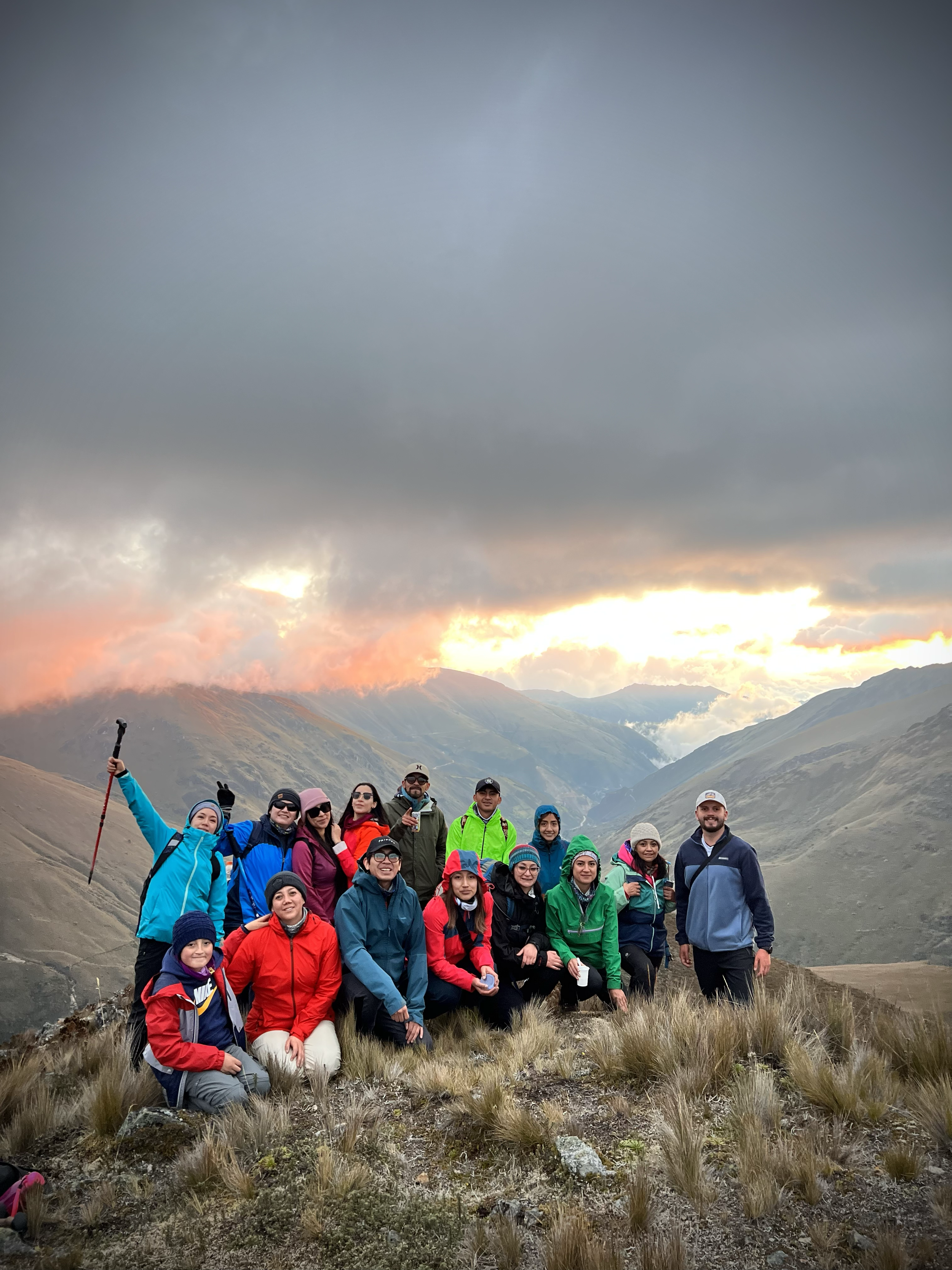 Grupo de personas en una cumbre de montaña con un paisaje montañoso y un cielo nublado y con luz de atardecer.