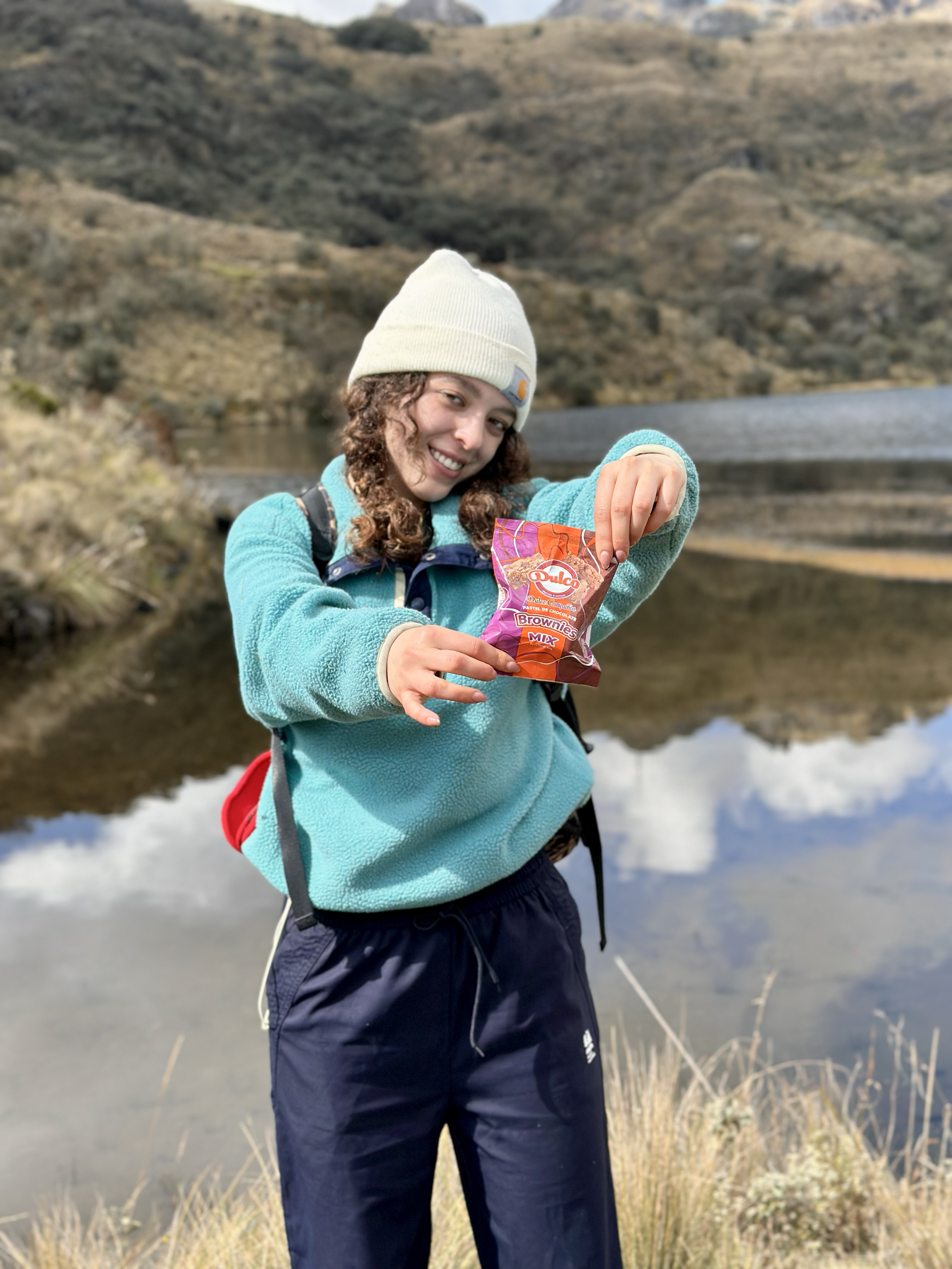 Una joven en ropa de senderismo sostiene una bolsa de brownies en un paisaje natural con lago y colinas en el fondo.