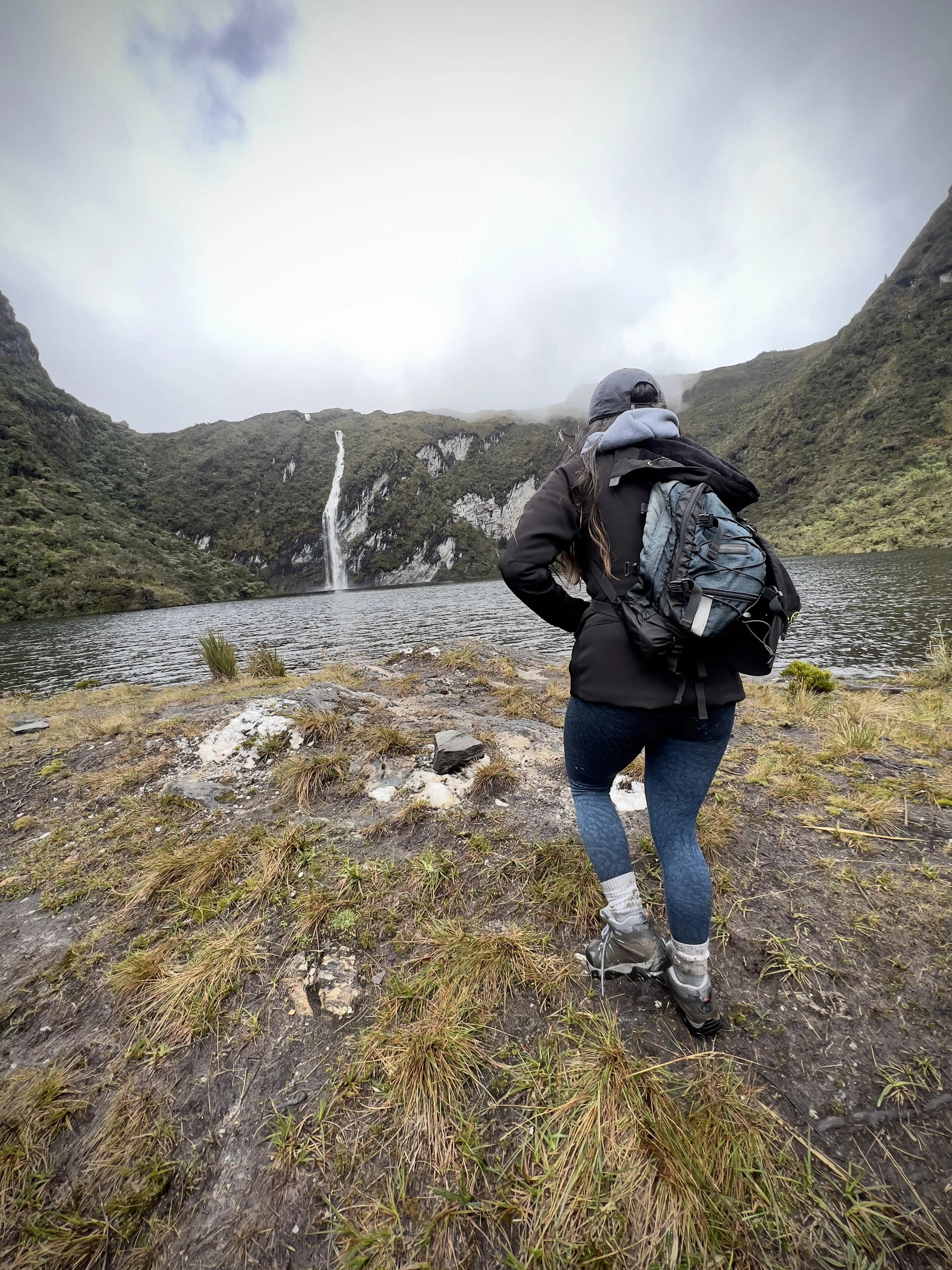 Una mujer con chaqueta y mochila observando cascada en un paisaje natural con laguna y montañas nubladas.