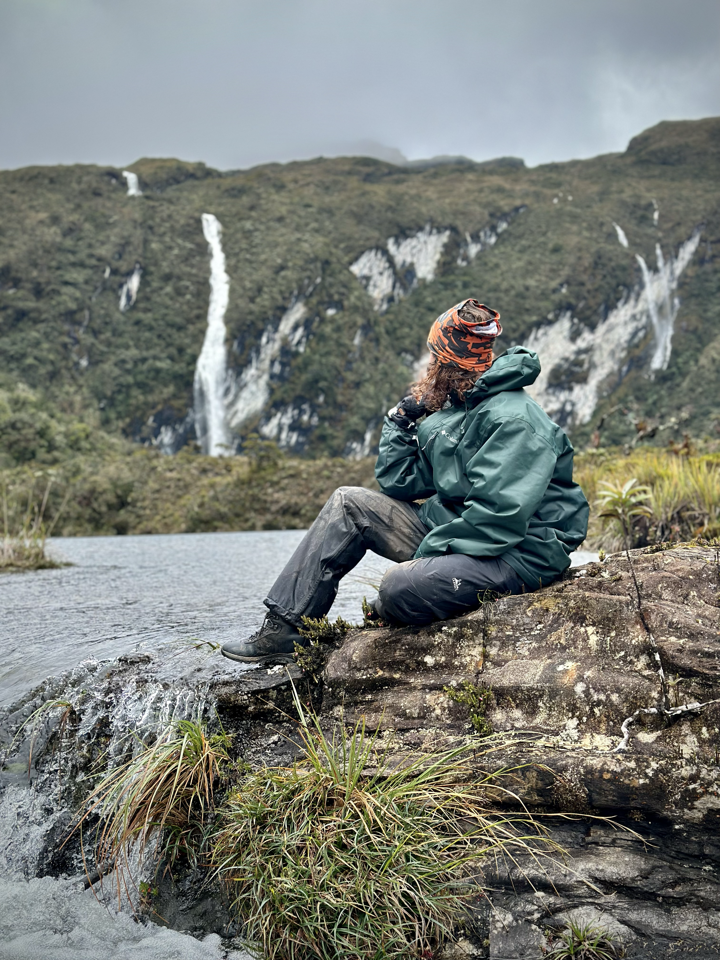 Persona sentada en una roca junto a un río, con montañas de fondo que tienen cascadas y vegetación, usando ropa de aventura y un gorro con estampado naranja y negro.