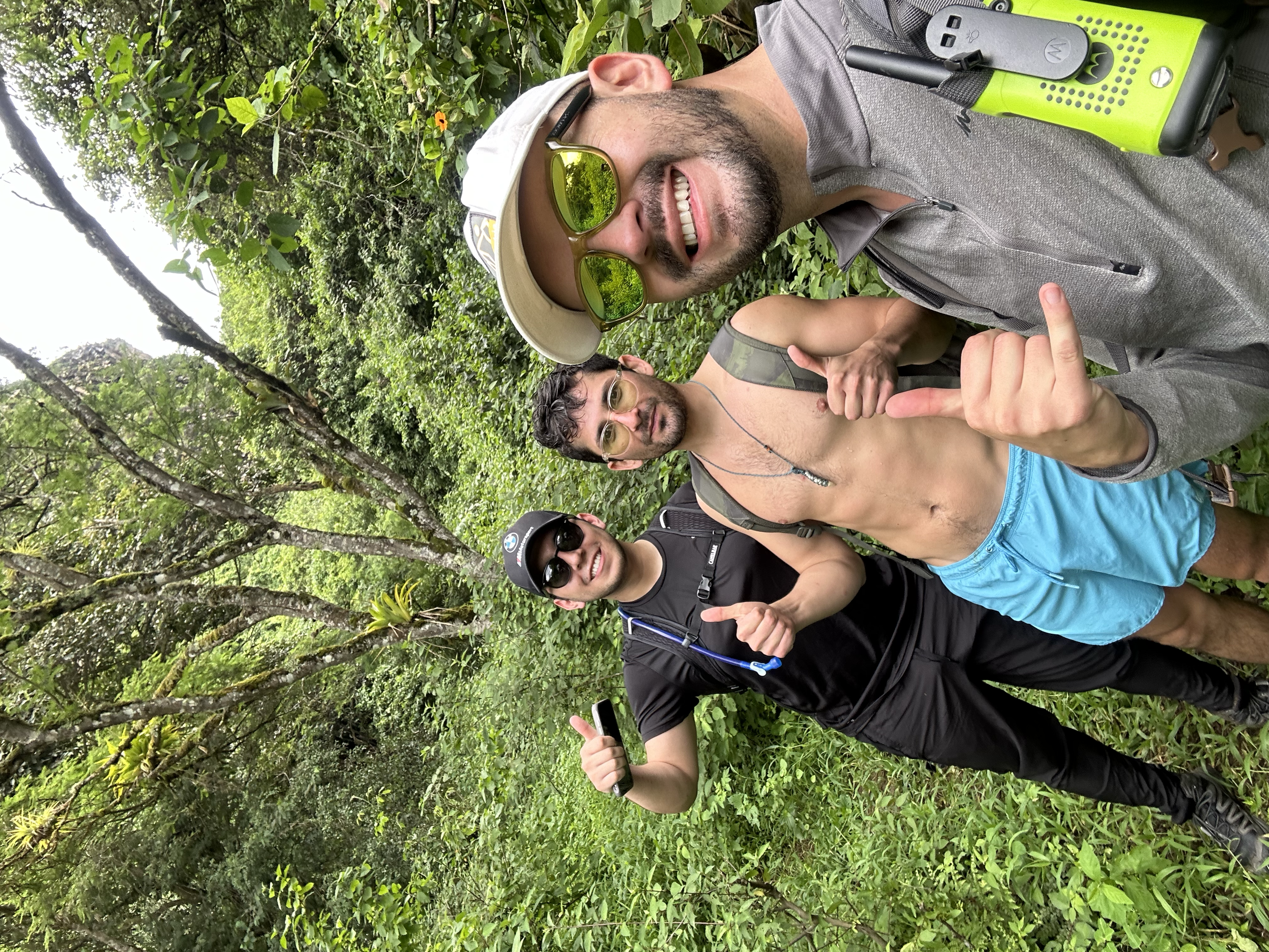 Tres hombres en un entorno de selva, sonriendo y tomando una selfie, uno de ellos con gafas de sol y otro sin camiseta, todos con mochilas y equipo de senderismo.