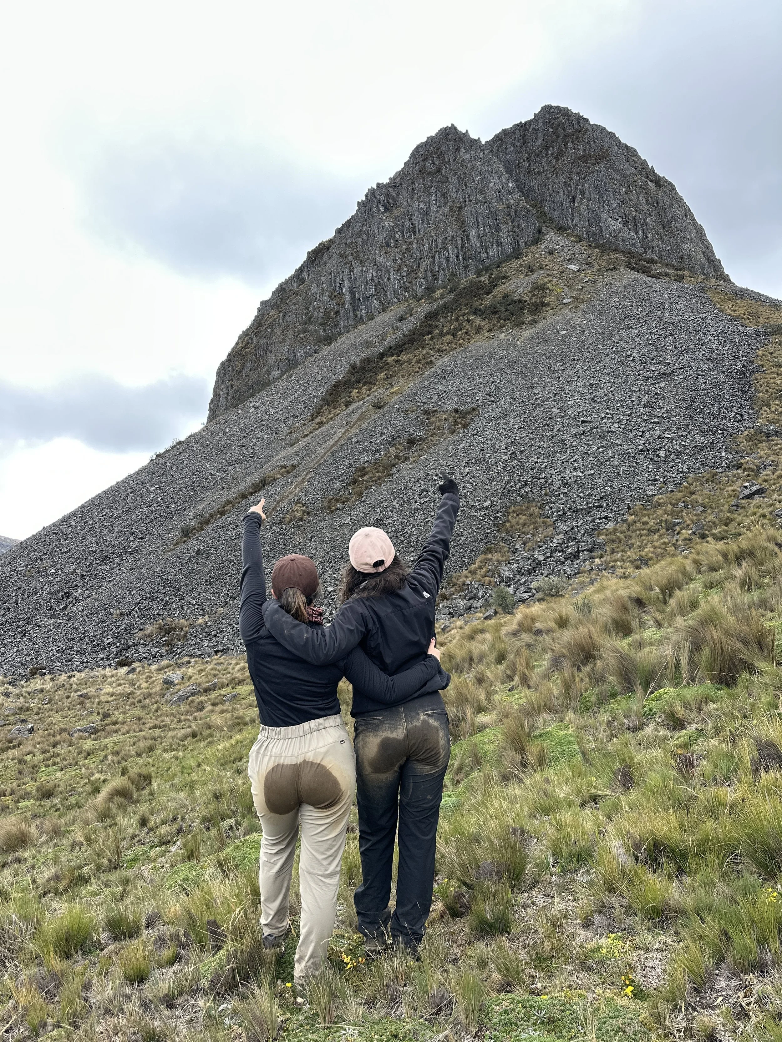 Dos personas de espaldas, abrazadas y señalando una montaña en un paisaje montañoso con césped y árboles, cielo nublado.