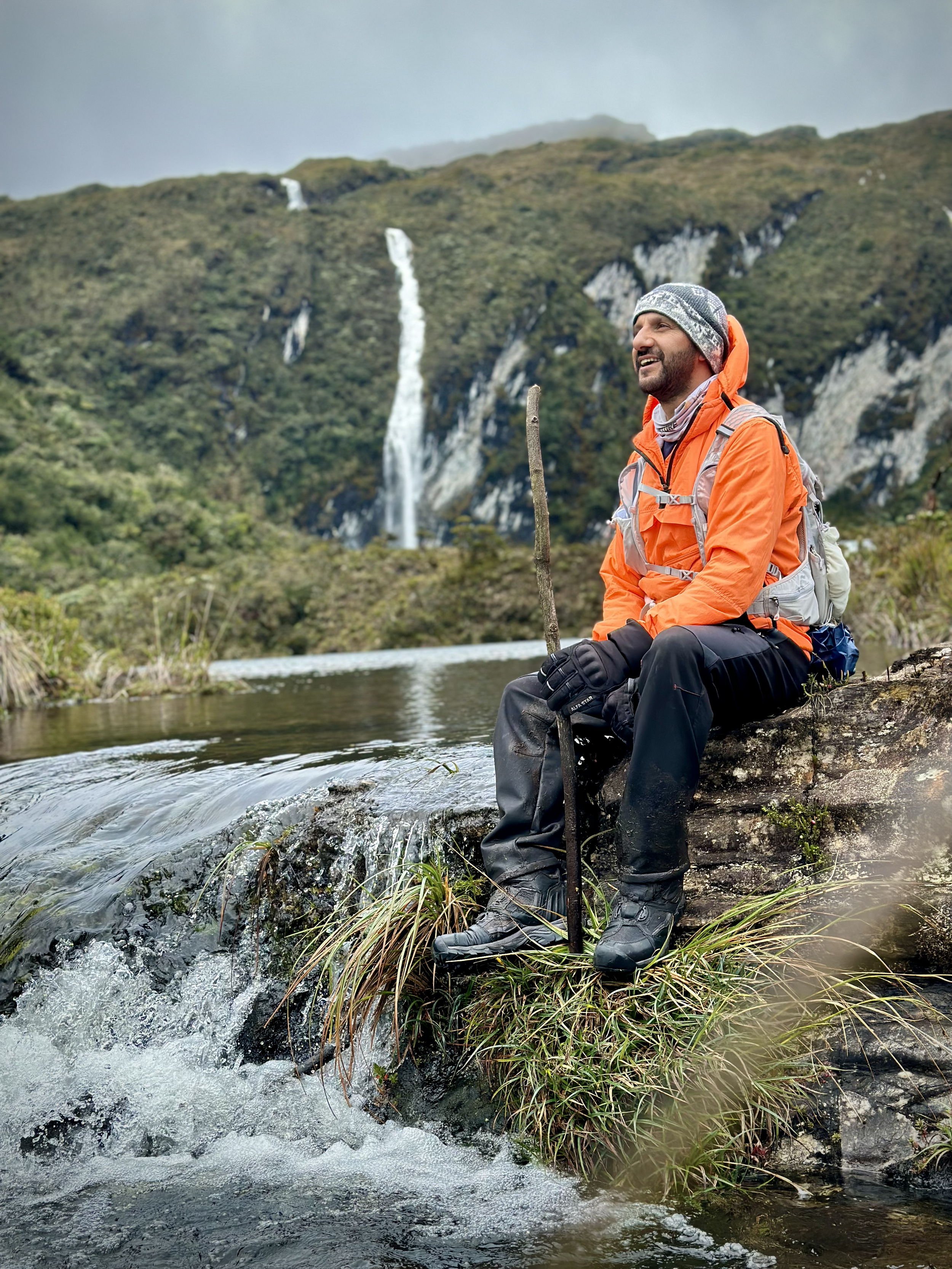 Hombre sentado junto a un río en un entorno de naturaleza con cascadas y montañas al fondo.